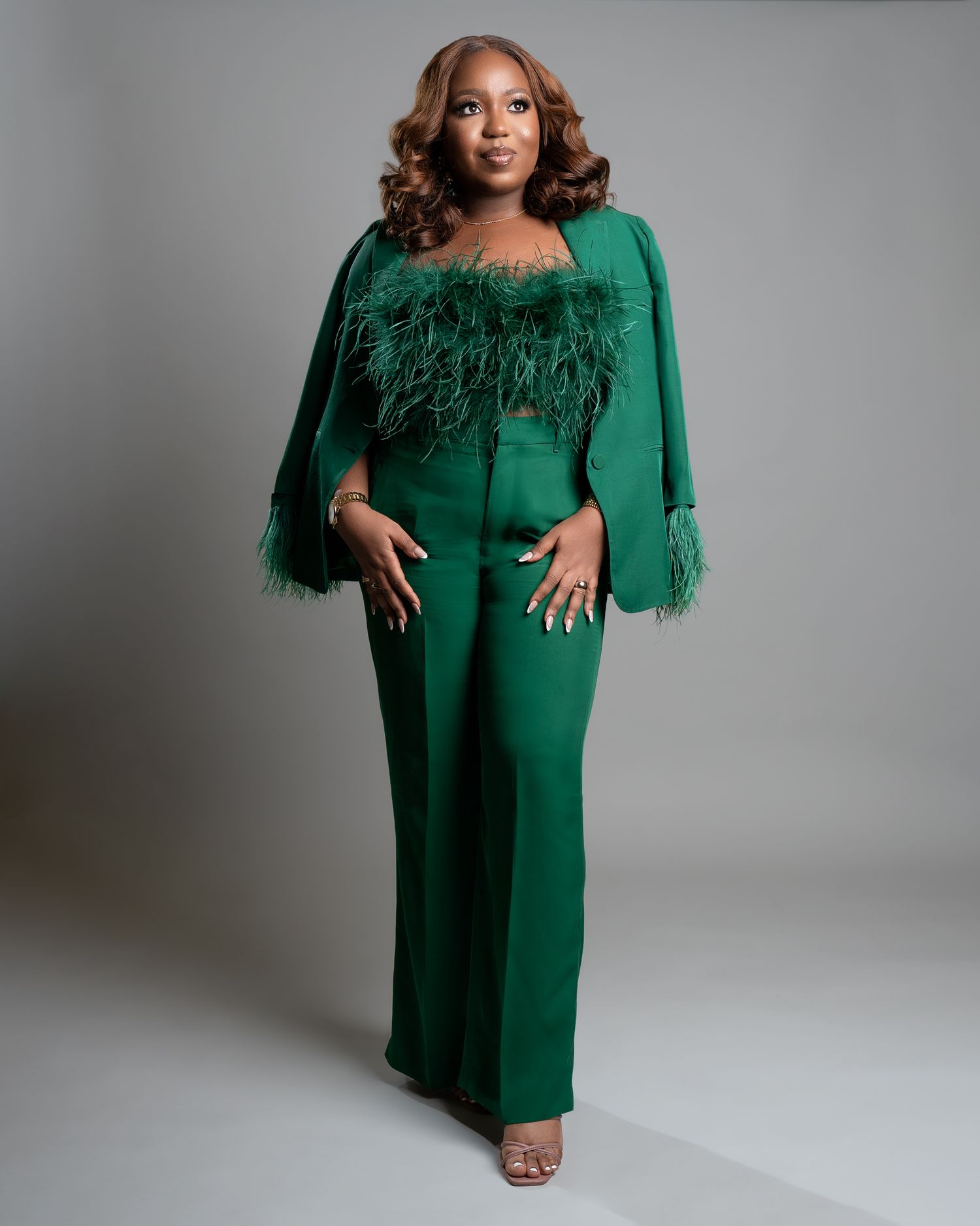 A figure poses in an elegant emerald green dress with feathered sleeves against a light gray studio backdrop.