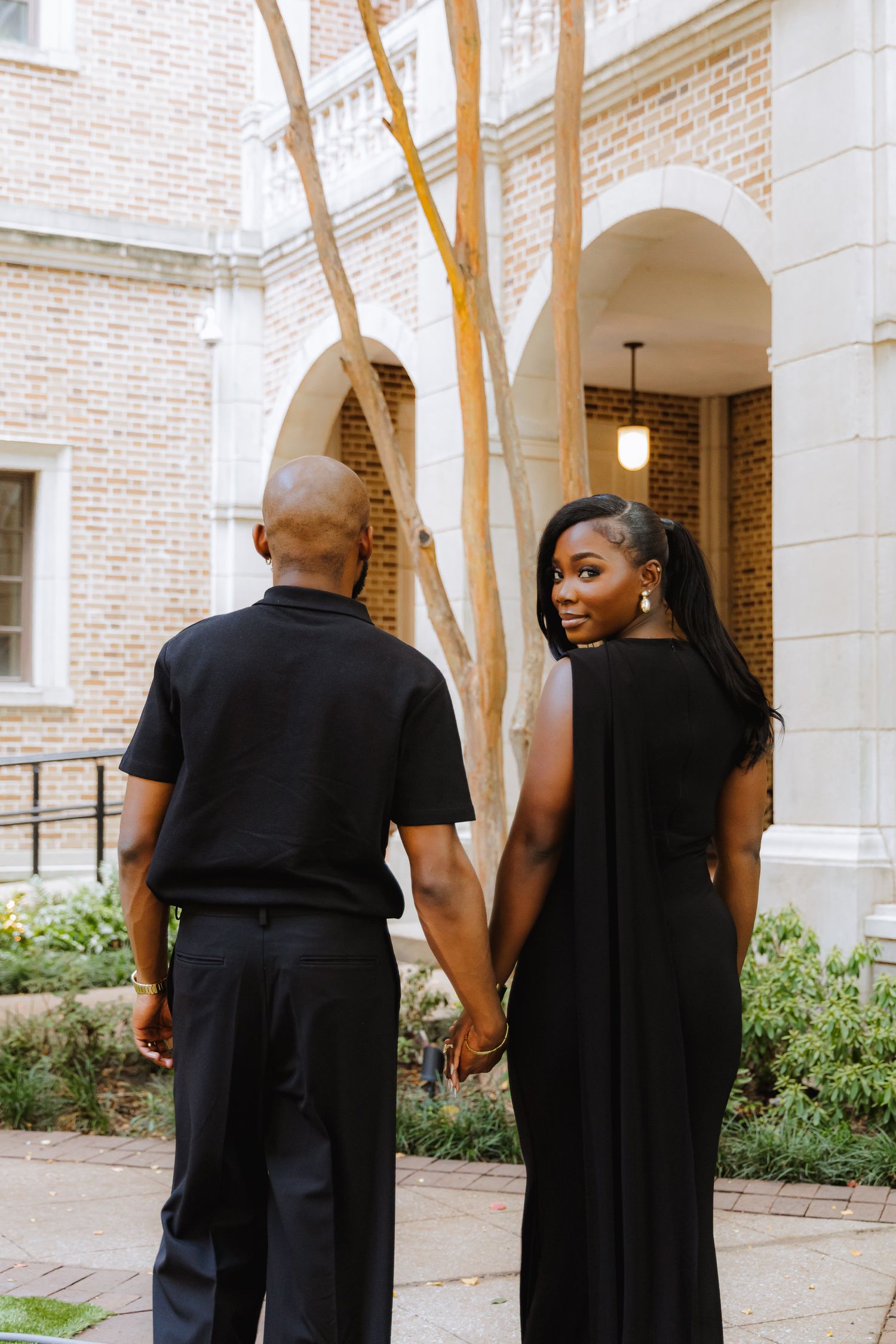Couple in matching black outfits walking together through a brick-columned courtyard.