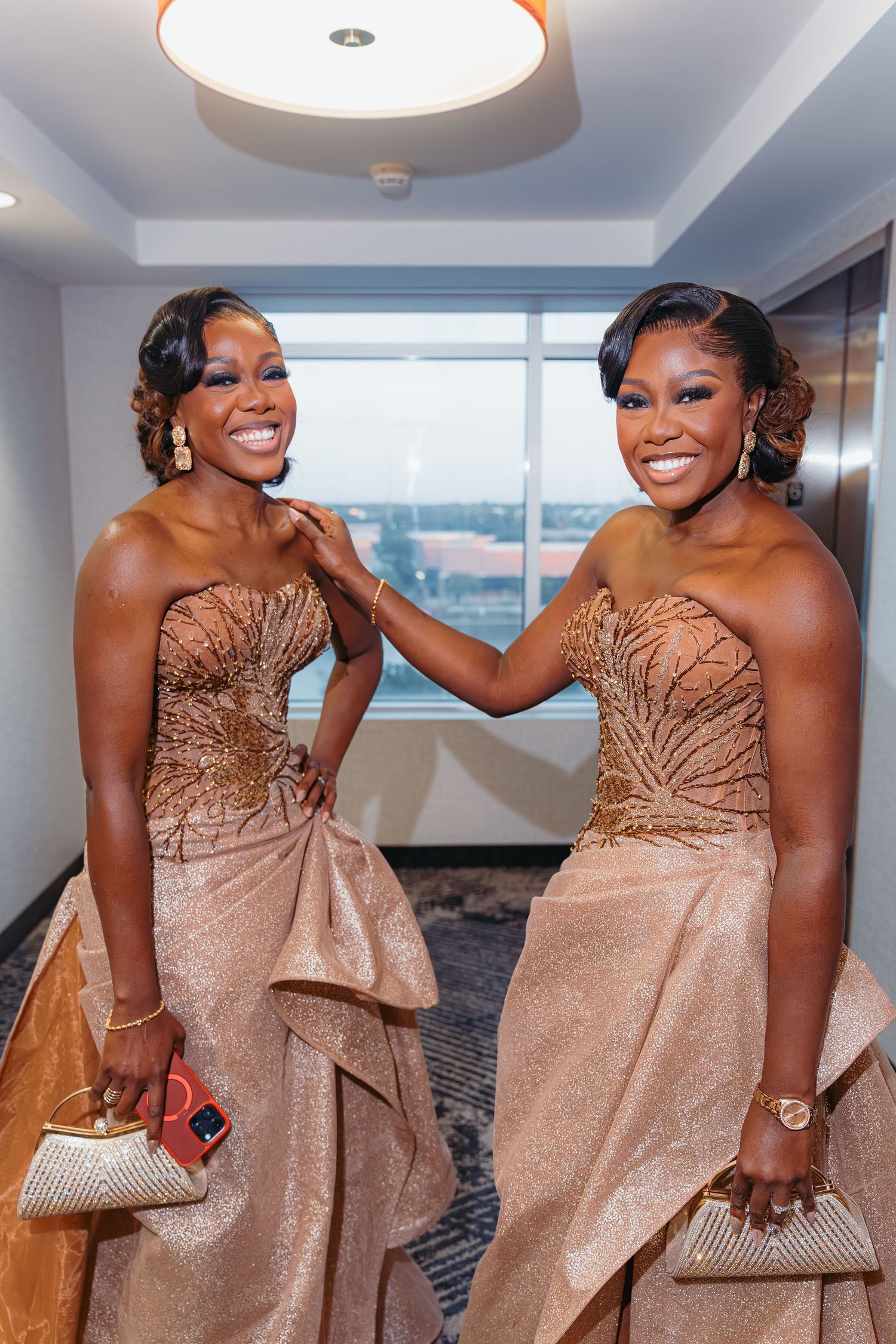 Two women wearing matching champagne-colored strapless gowns with beaded details pose together in an elegant interior setting.