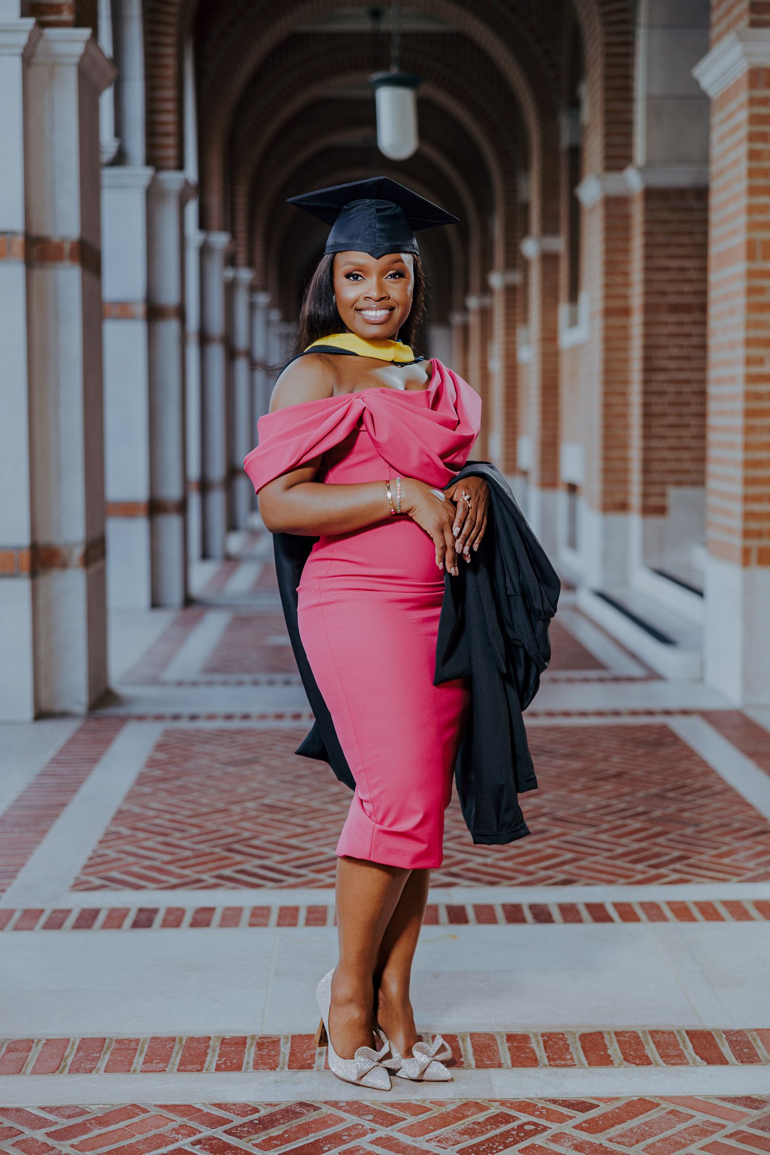 Graduate in pink off-shoulder dress and cap poses in arched hallway with brick columns and tiled floor.