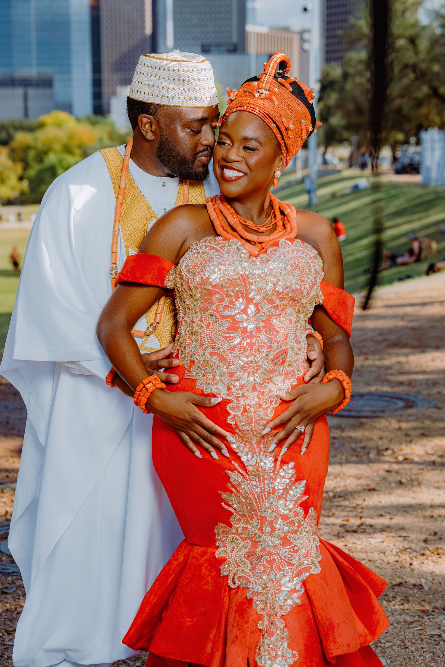 A couple in traditional African wedding attire with red and white colors embracing outdoors in a park setting.