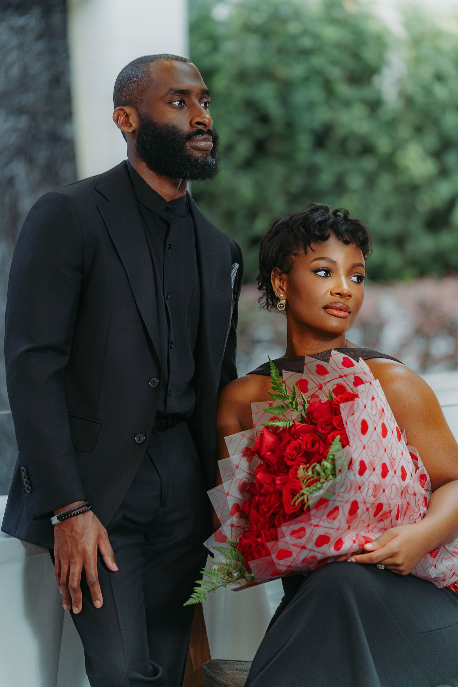 A romantic couple in formal black and red attire pose together with red roses and heart-themed decor for engagement photos.