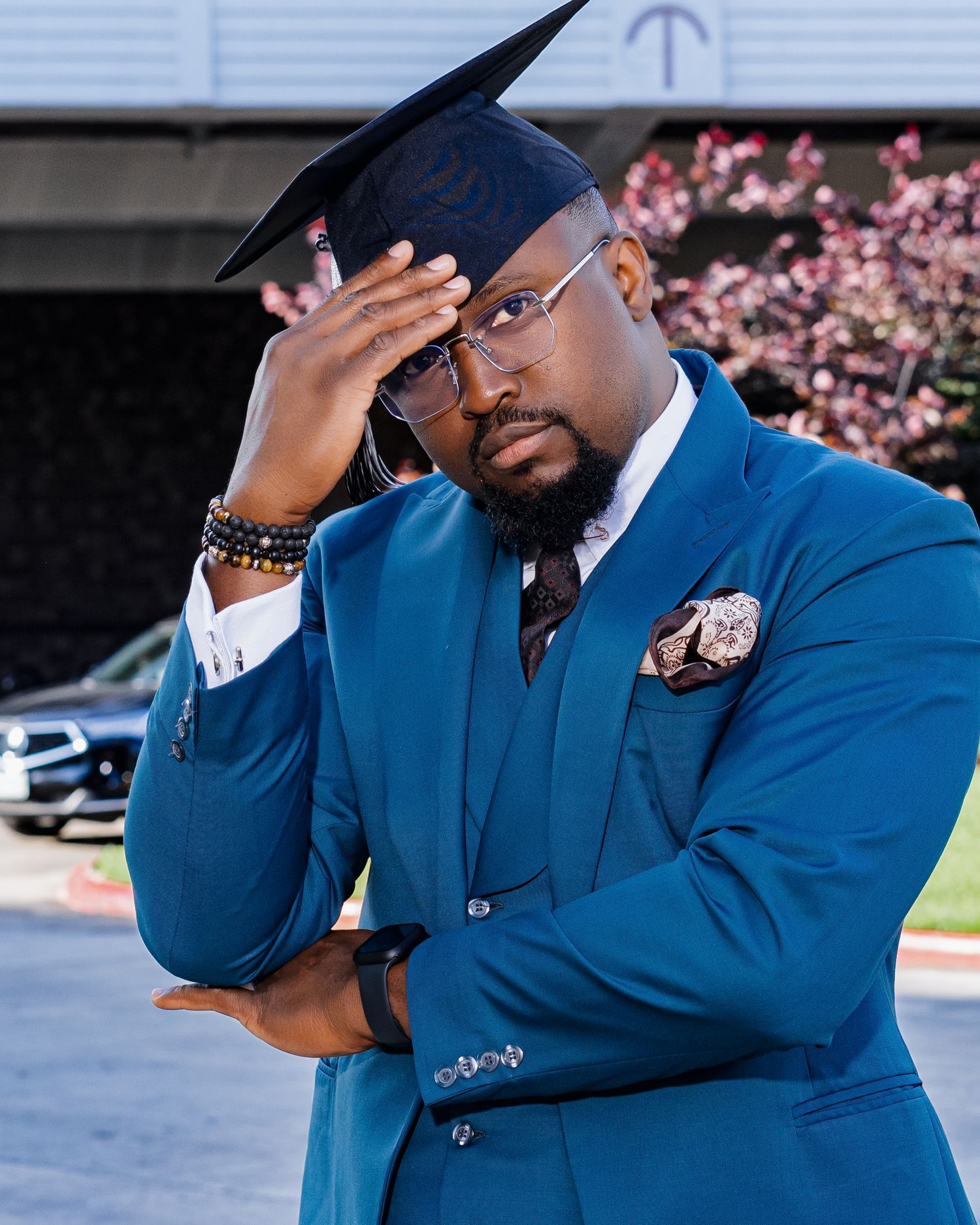 A figure in a blue suit and hat poses thoughtfully against a backdrop of pink cherry blossoms.