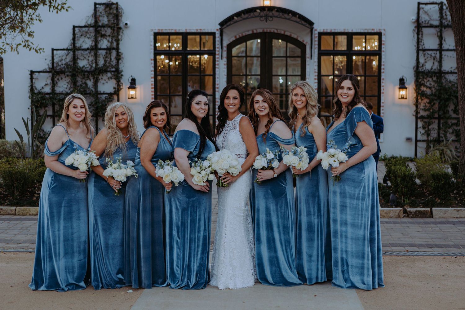 Wedding party in white and blue dresses posing outside elegant venue.