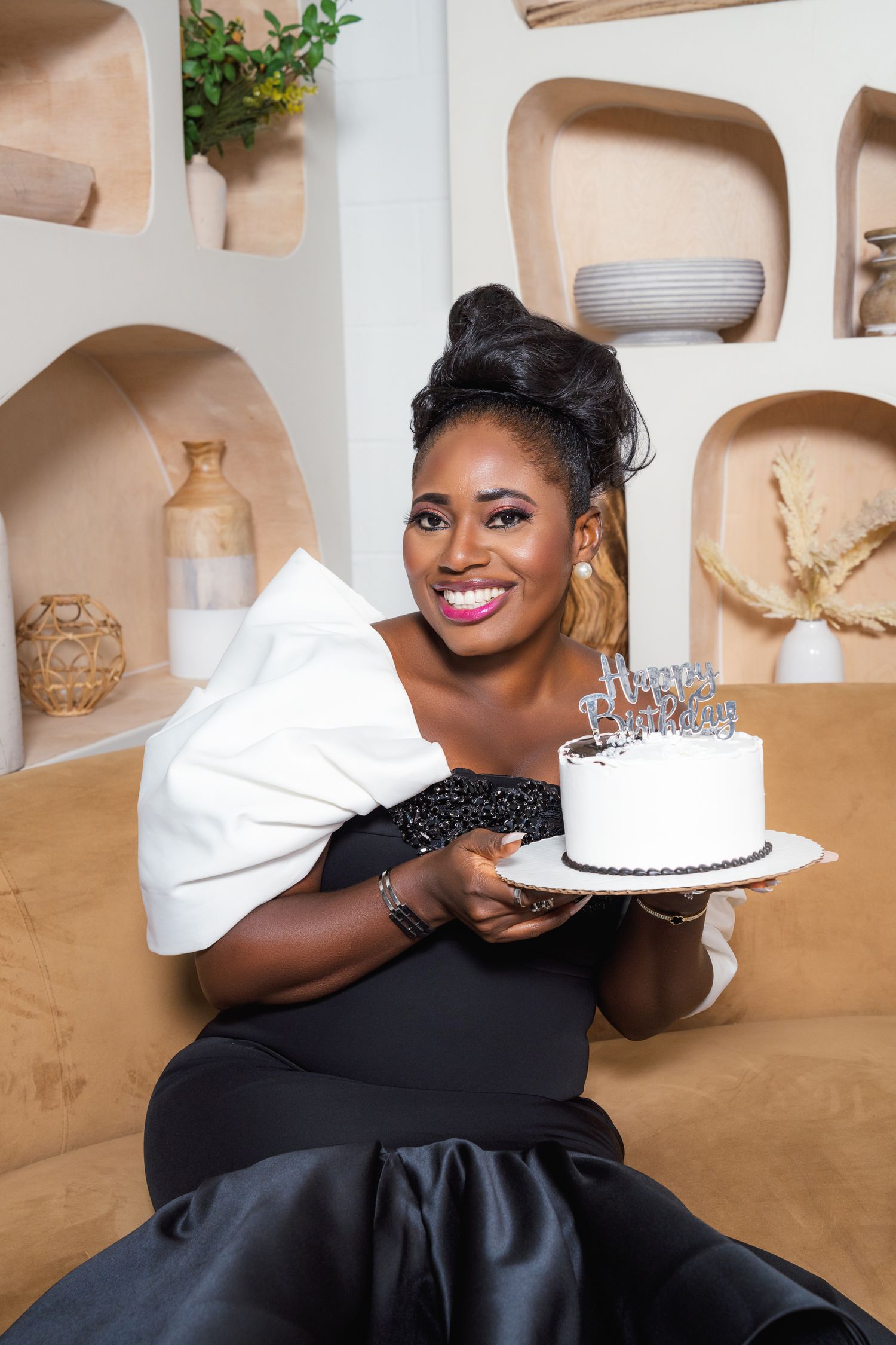 A person in an elegant black evening gown poses with a decorative cake against a beige background.