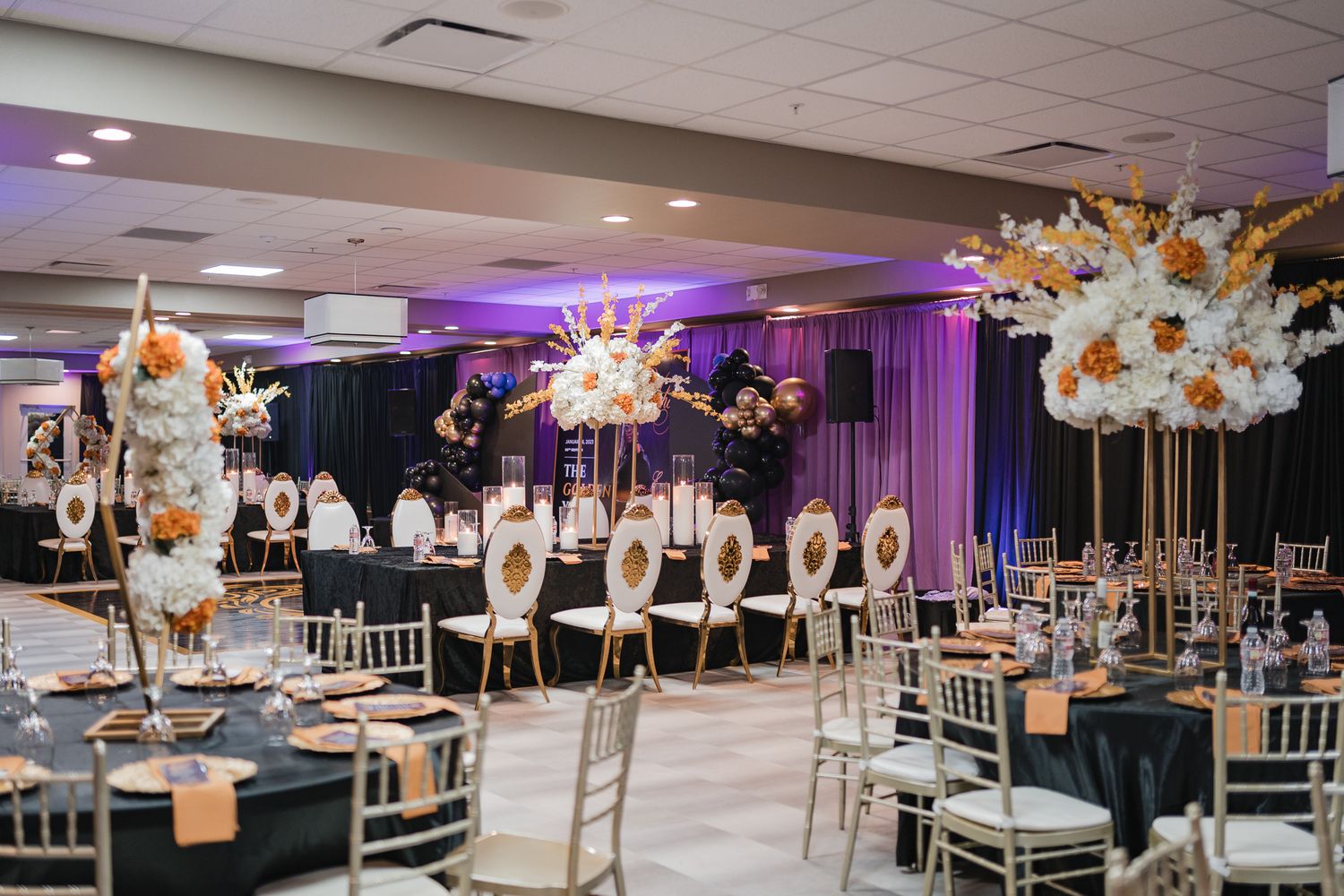 Elegant ballroom setup with black and gold decor, tall floral centerpieces, and purple uplighting for a formal event.