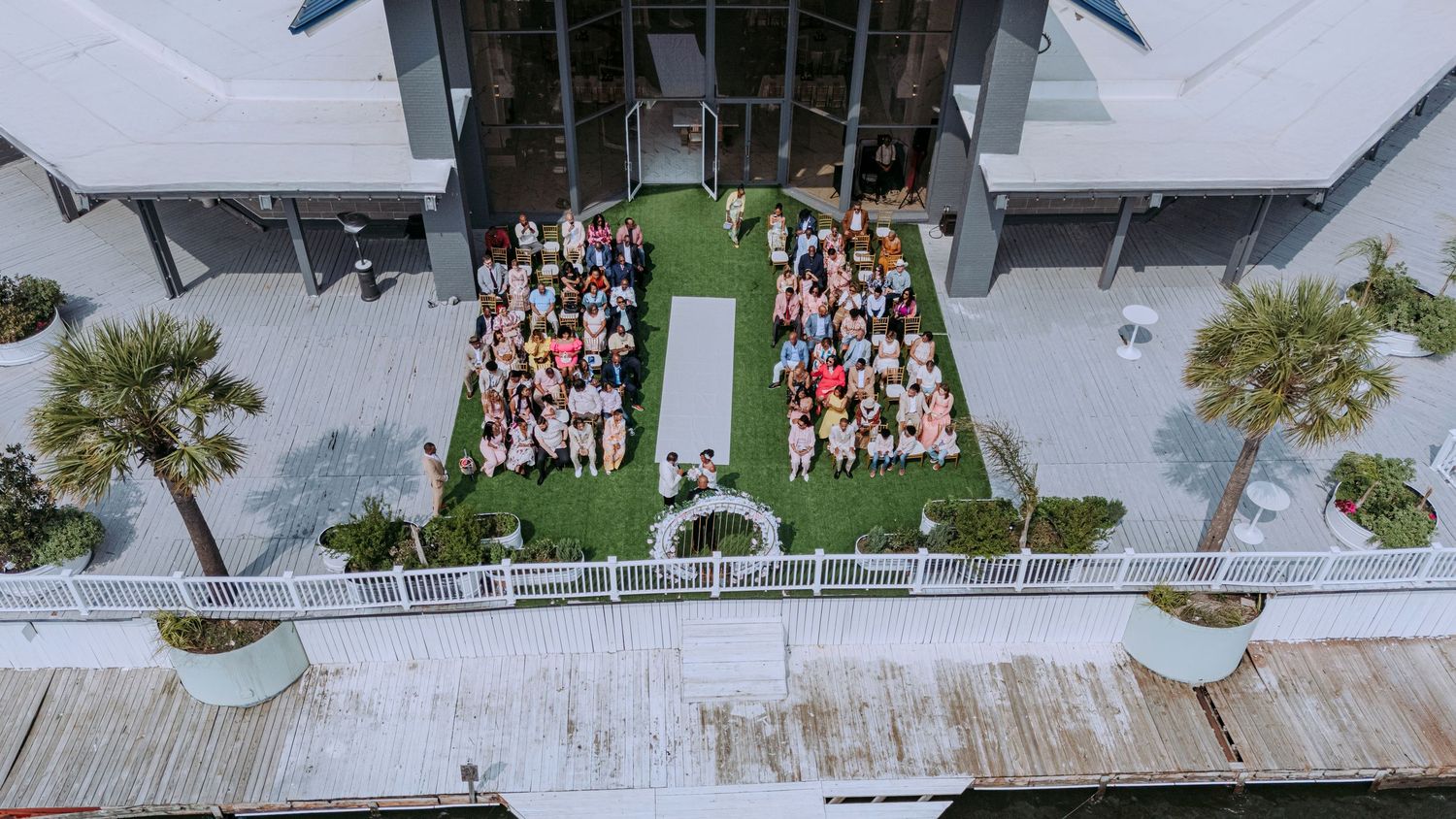 Aerial view of a beachfront wedding ceremony setup with white chairs and palm trees.