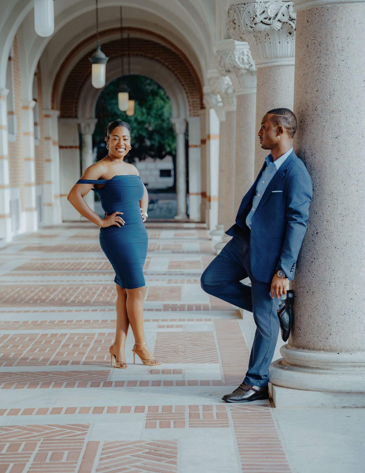 Couple in coordinating blue outfits posing in elegant columned hallway, woman in off-shoulder dress, man in suit leaning on pillar.