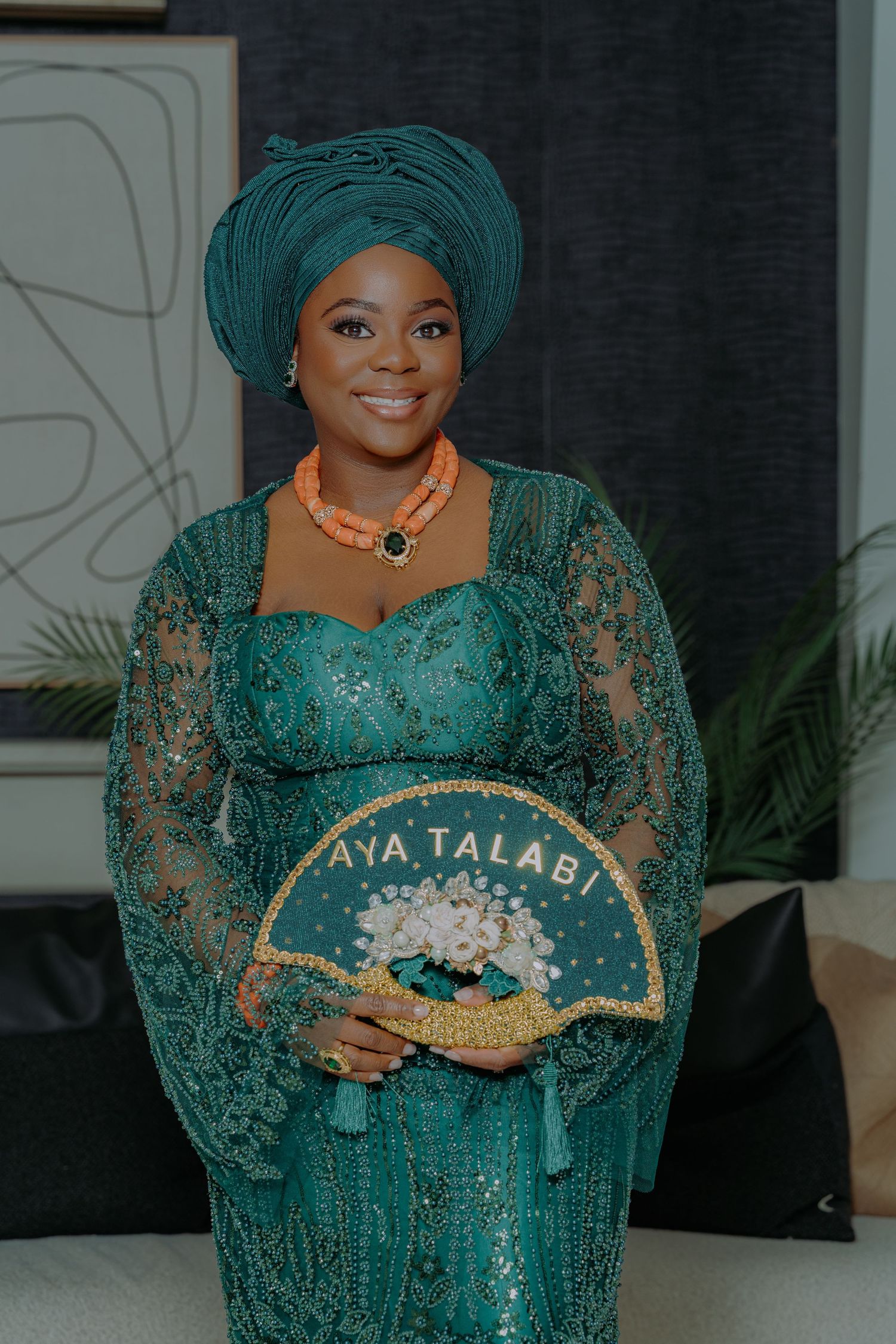 Woman in teal Nigerian lace gown and gele headwrap holds decorative fan reading Aya Talabi at traditional Yoruba ceremony.