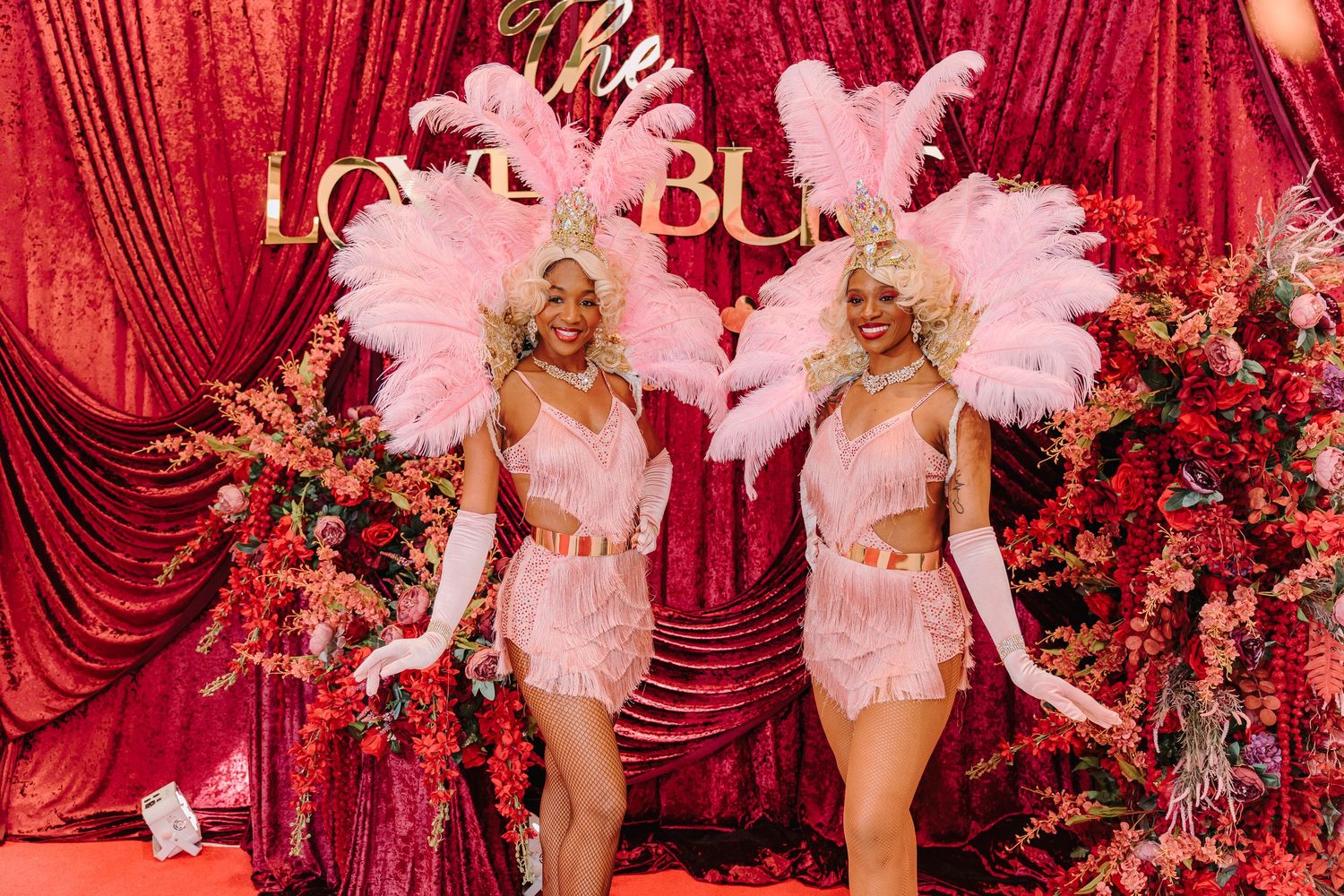 Showgirls in pink feathered costumes perform on stage against red curtains and floral decorations.