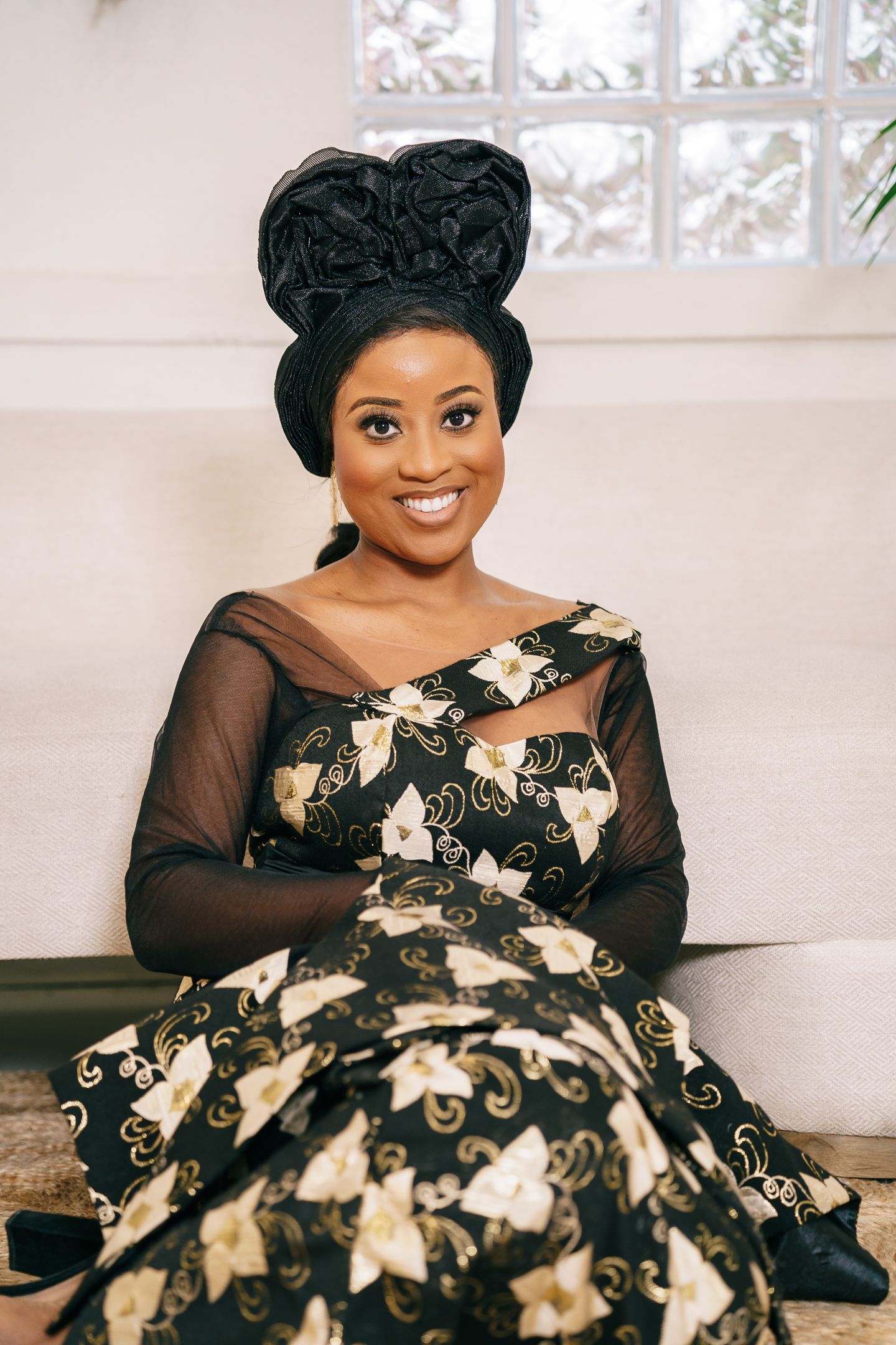 A person in an elegant black and gold evening gown with sheer sleeves poses on a white couch.
