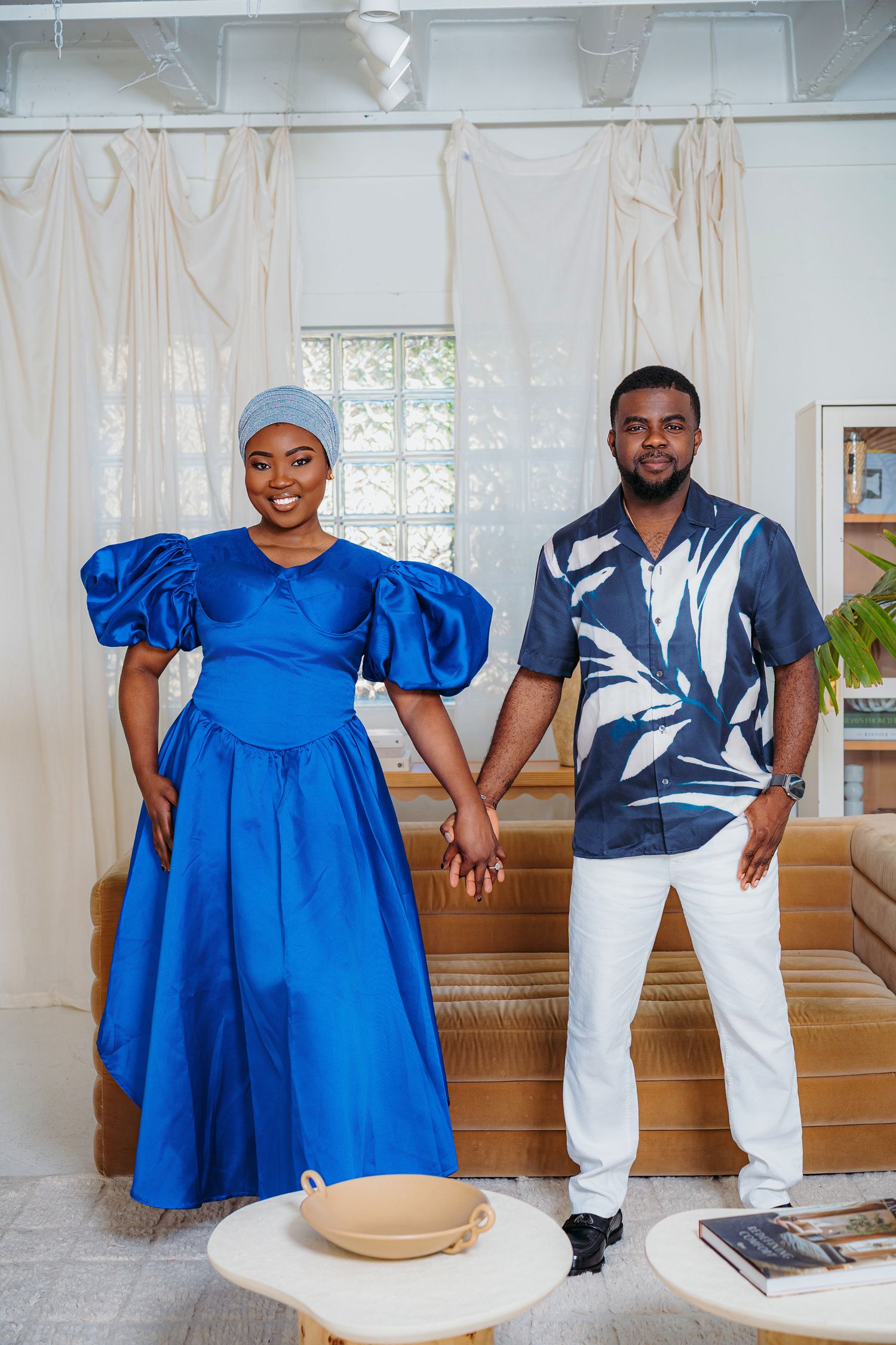Couple in coordinating blue outfits holding hands, woman in puff-sleeve gown, man in floral shirt, indoors.