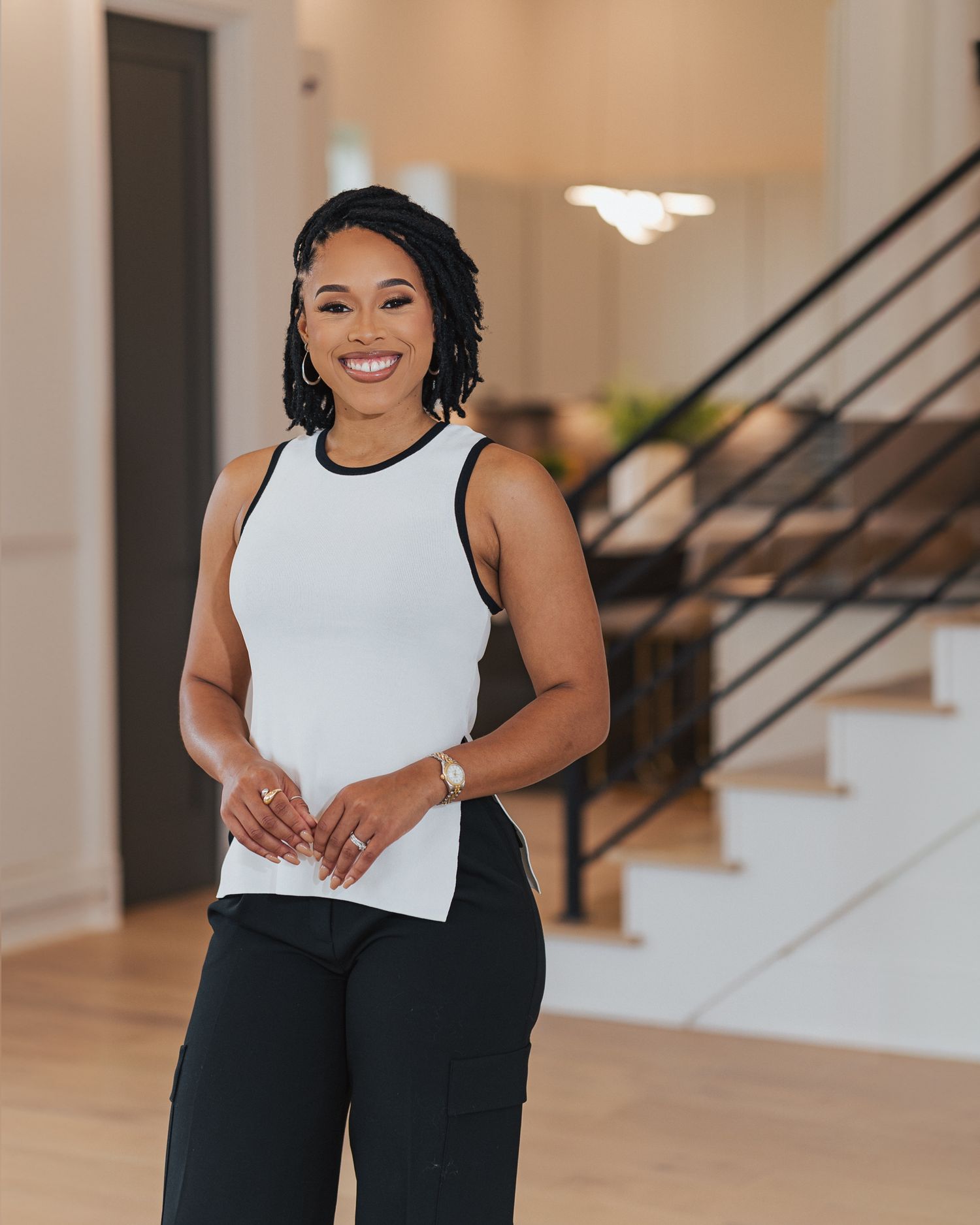 A person in a white sleeveless top and black pants smiles warmly while standing in a modern home interior.