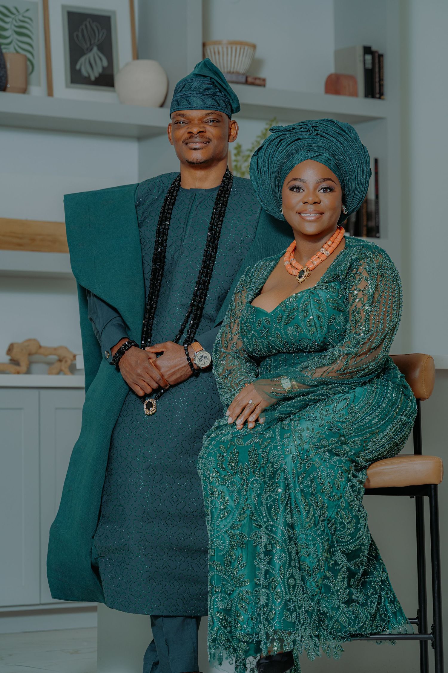 Nigerian couple in matching teal traditional Yoruba attire, agbada and lace gele, posing elegantly indoors.