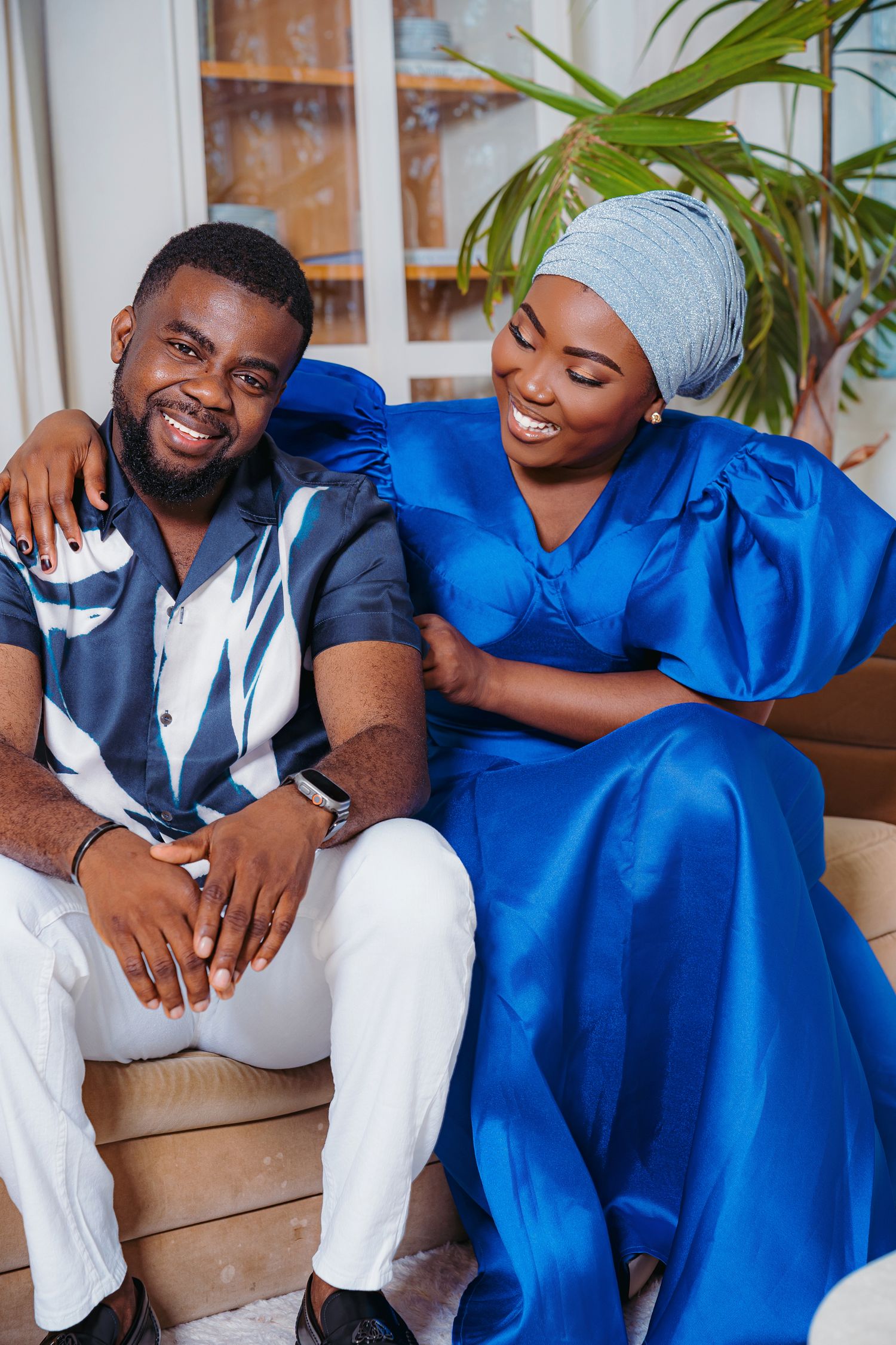 A smiling couple in blue outfits sit together on a sofa, the woman wearing a blue gele headwrap and satin dress.