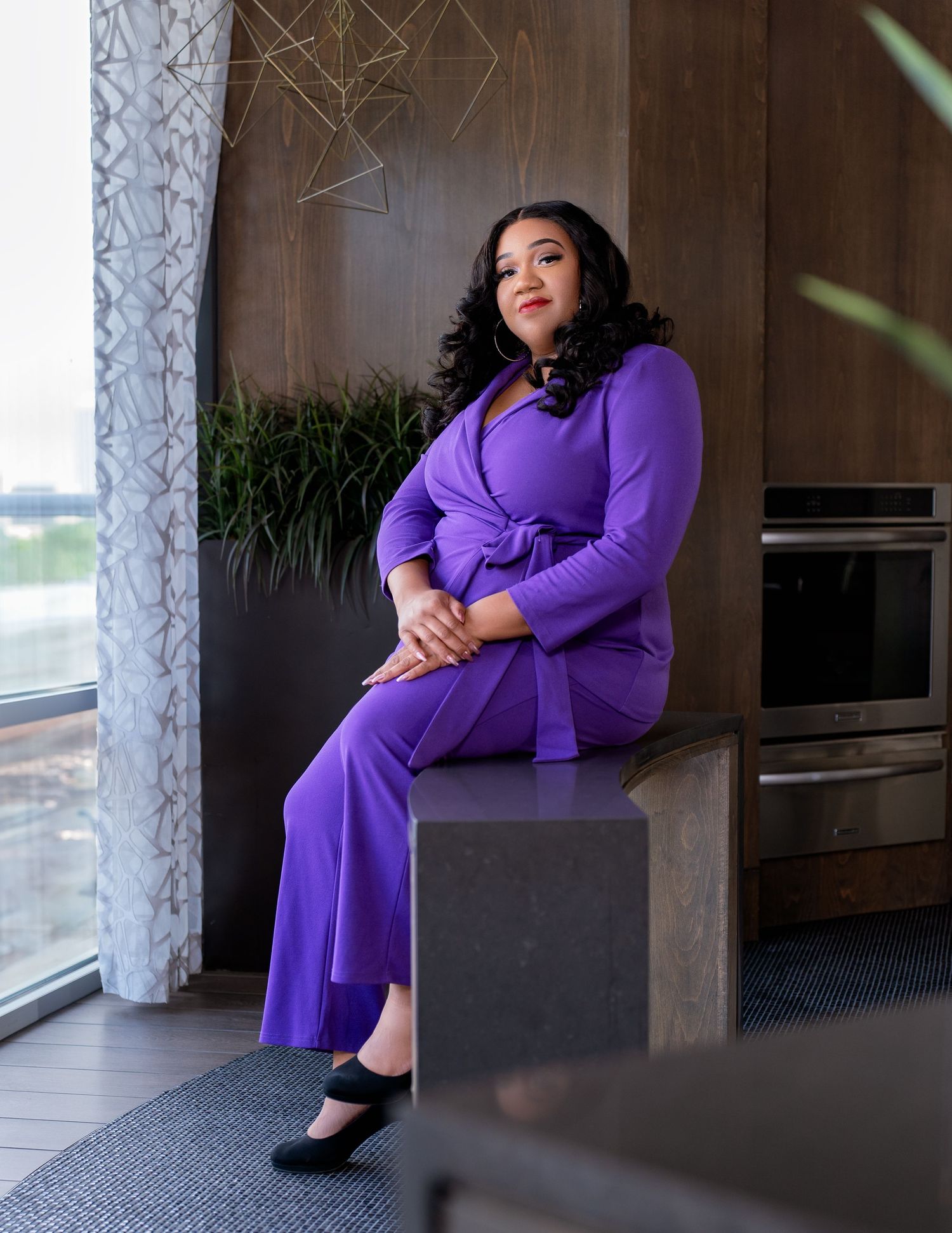 Woman in purple jumpsuit sitting on a stone pedestal in a modern kitchen interior.