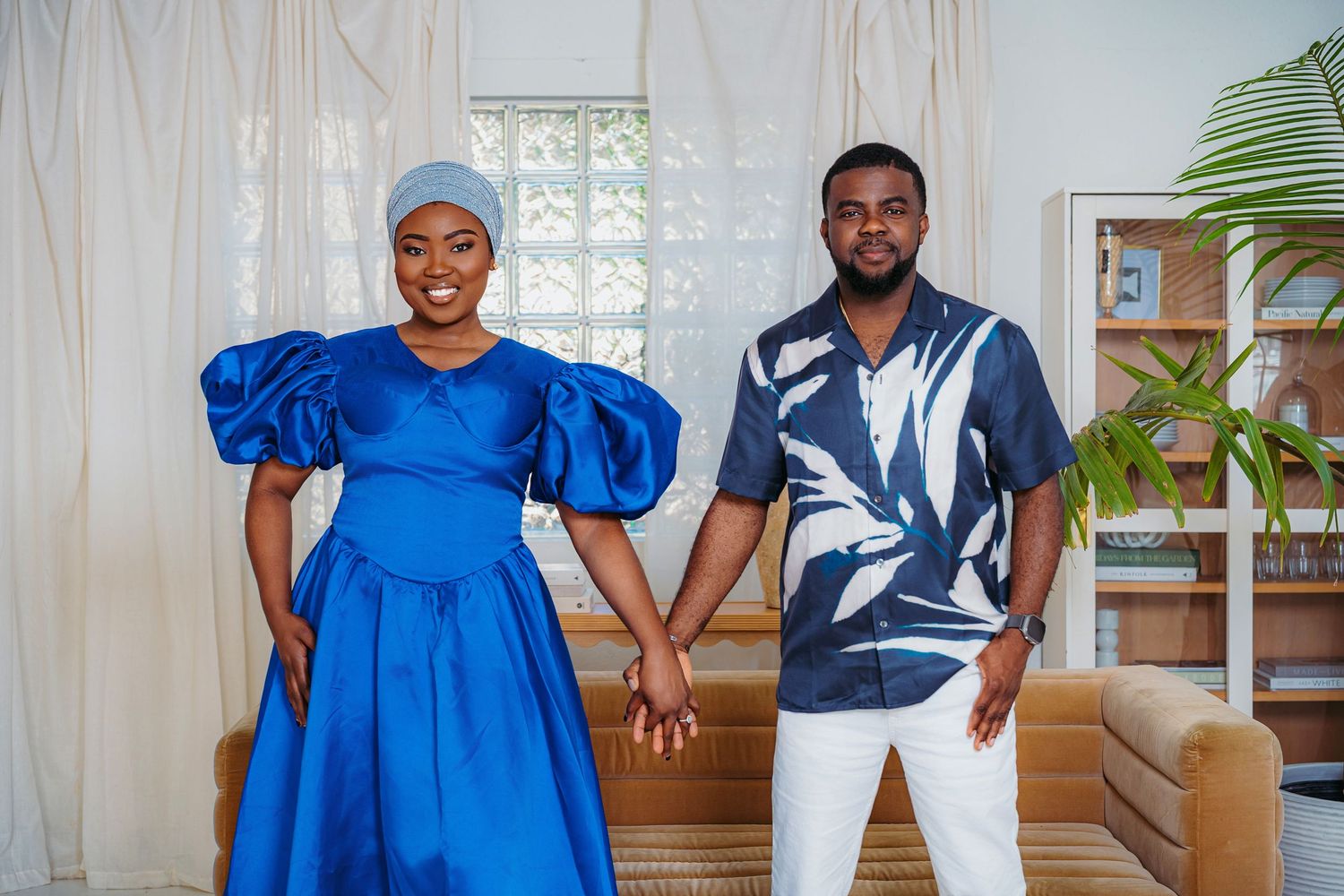 A couple in coordinating blue outfits hold hands and smile while posing indoors in a bright, elegant room.