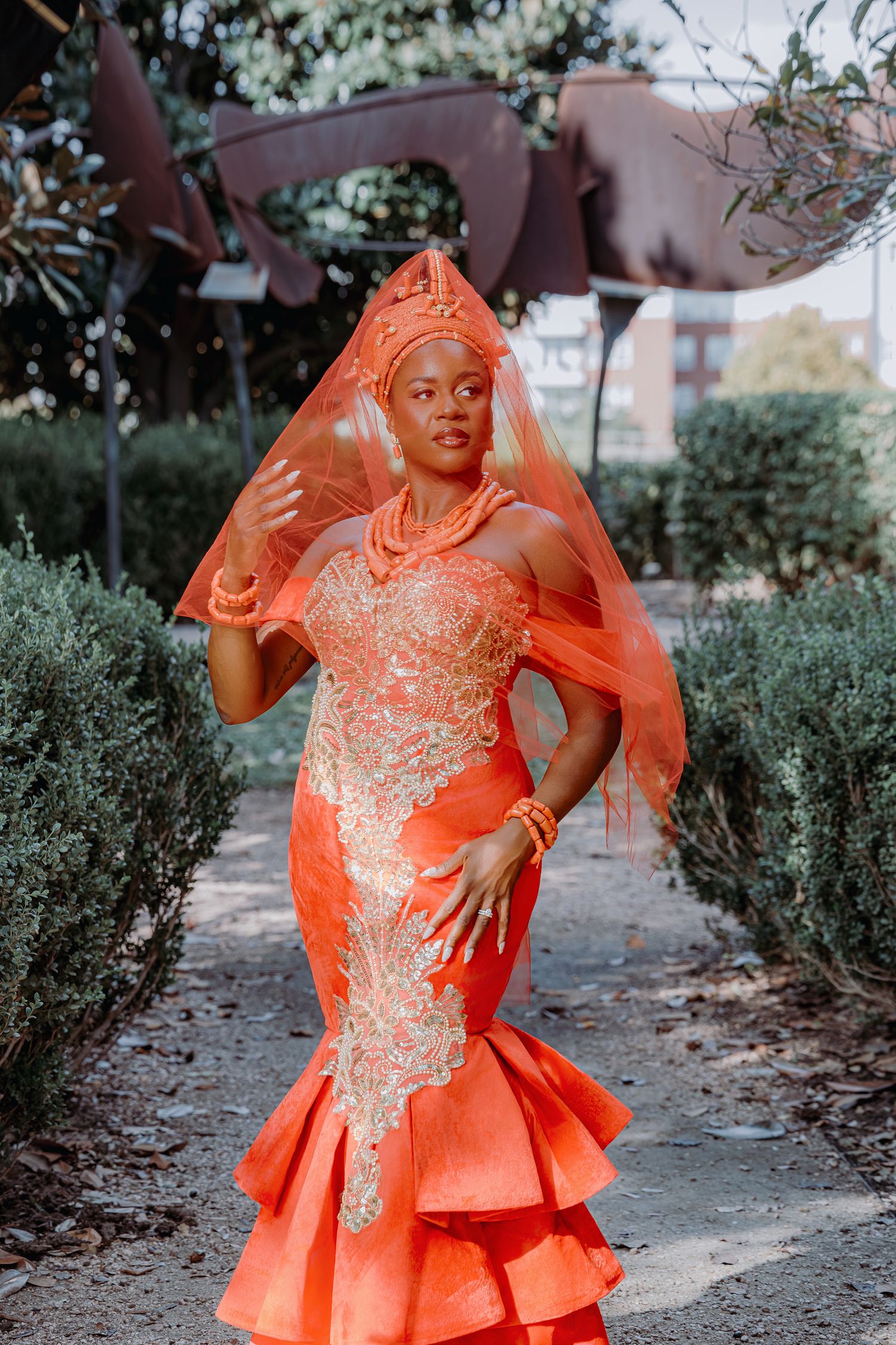 A person in a vibrant orange formal dress with intricate beading poses gracefully in a garden setting.