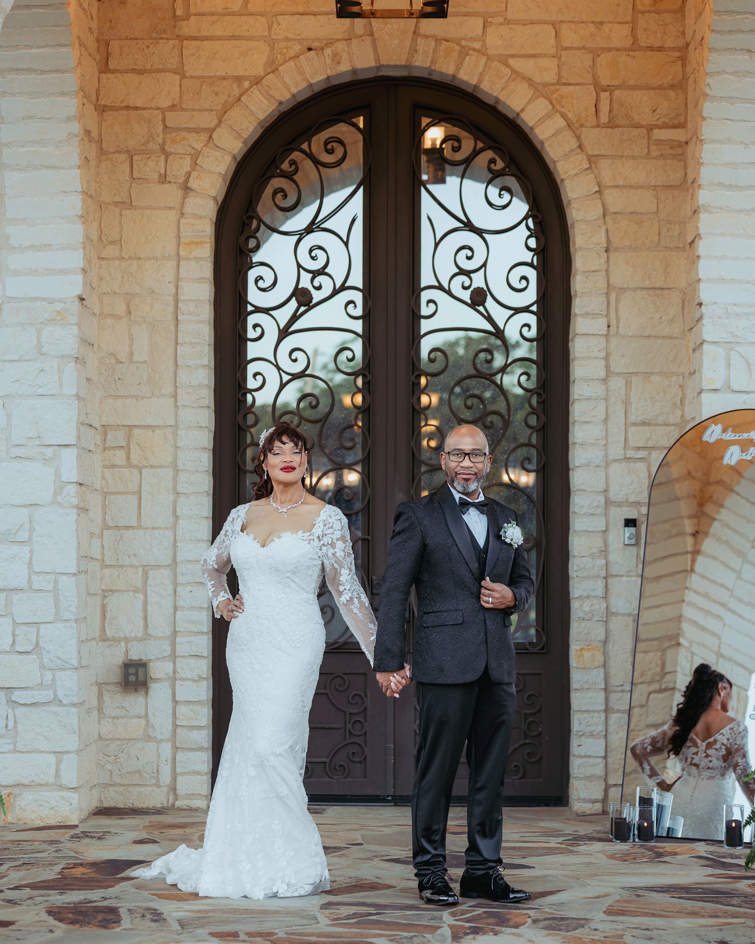 Newlyweds holding hands in front of ornate stone archway with decorative iron doors.