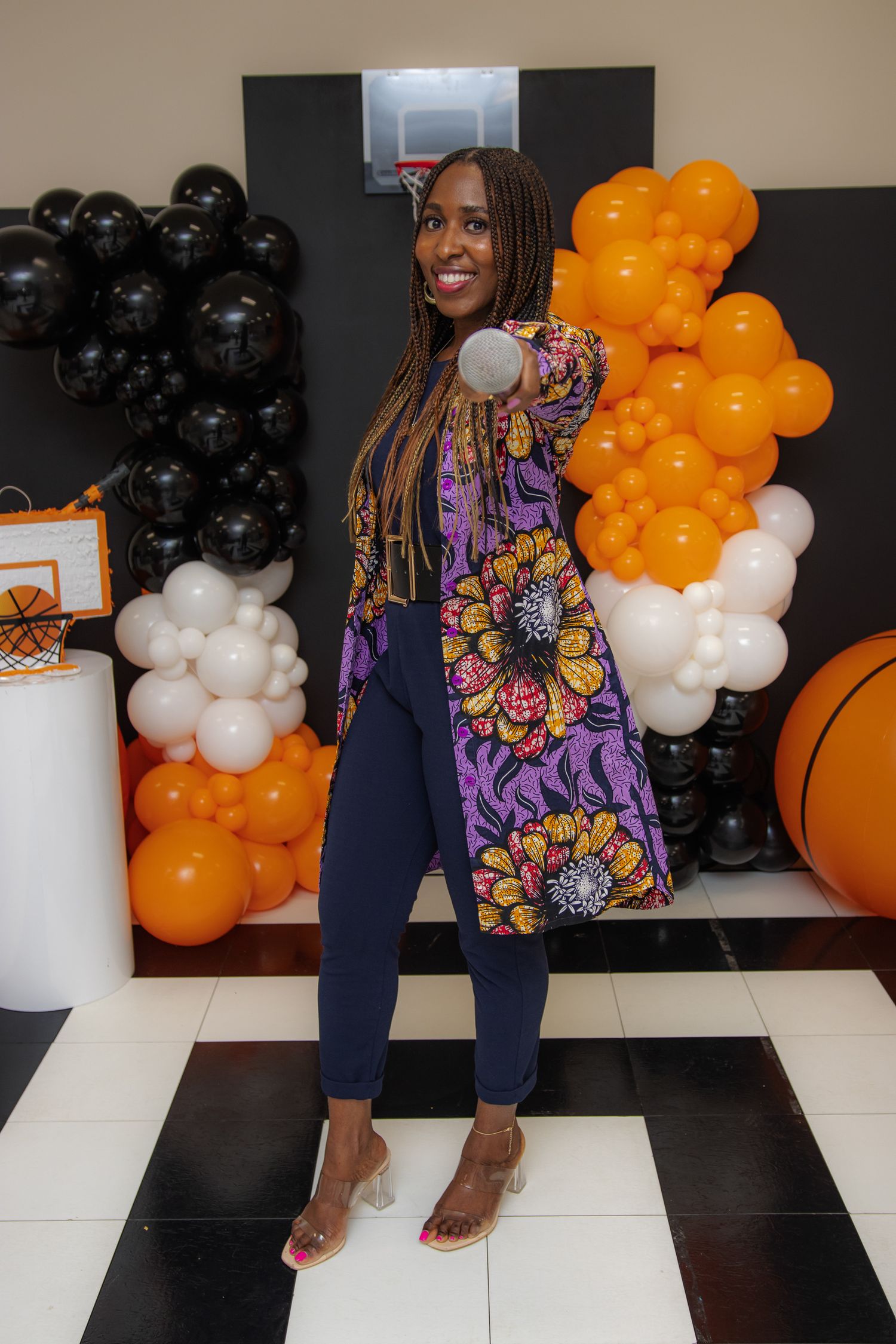 Woman in floral jacket poses with basketball at orange and black balloon party decoration backdrop.