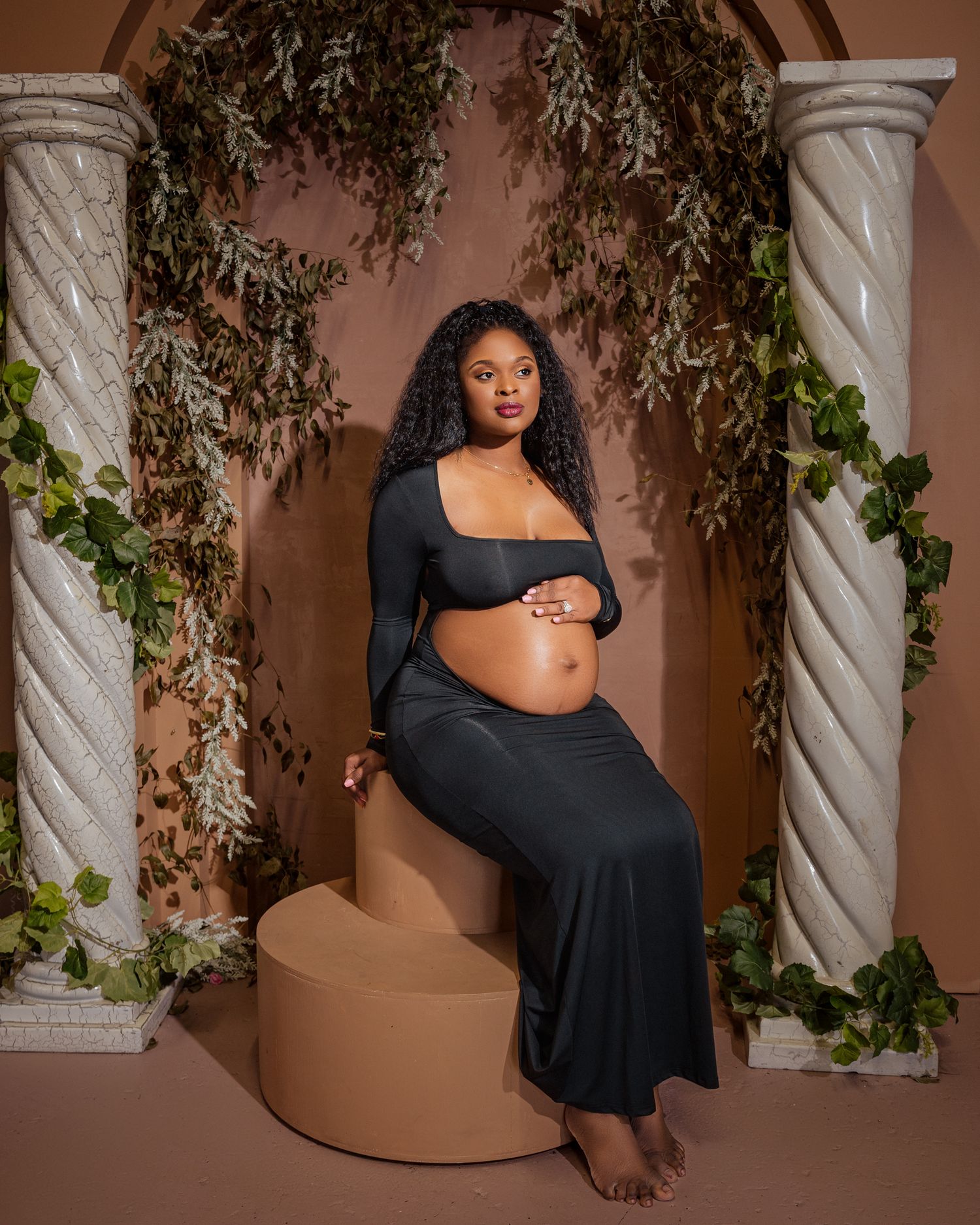 Maternity photoshoot in a romantic setting with spiral columns, draping greenery, and warm lighting against a terracotta backdrop.