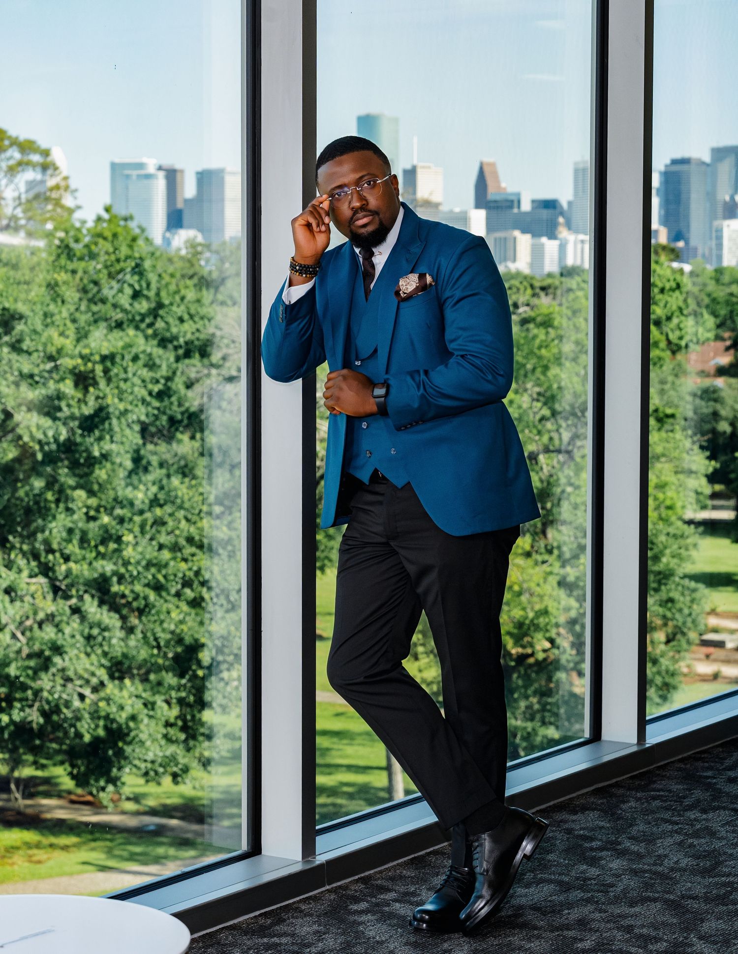 Professional in a blue blazer and dark pants poses thoughtfully by large windows overlooking a green urban landscape.