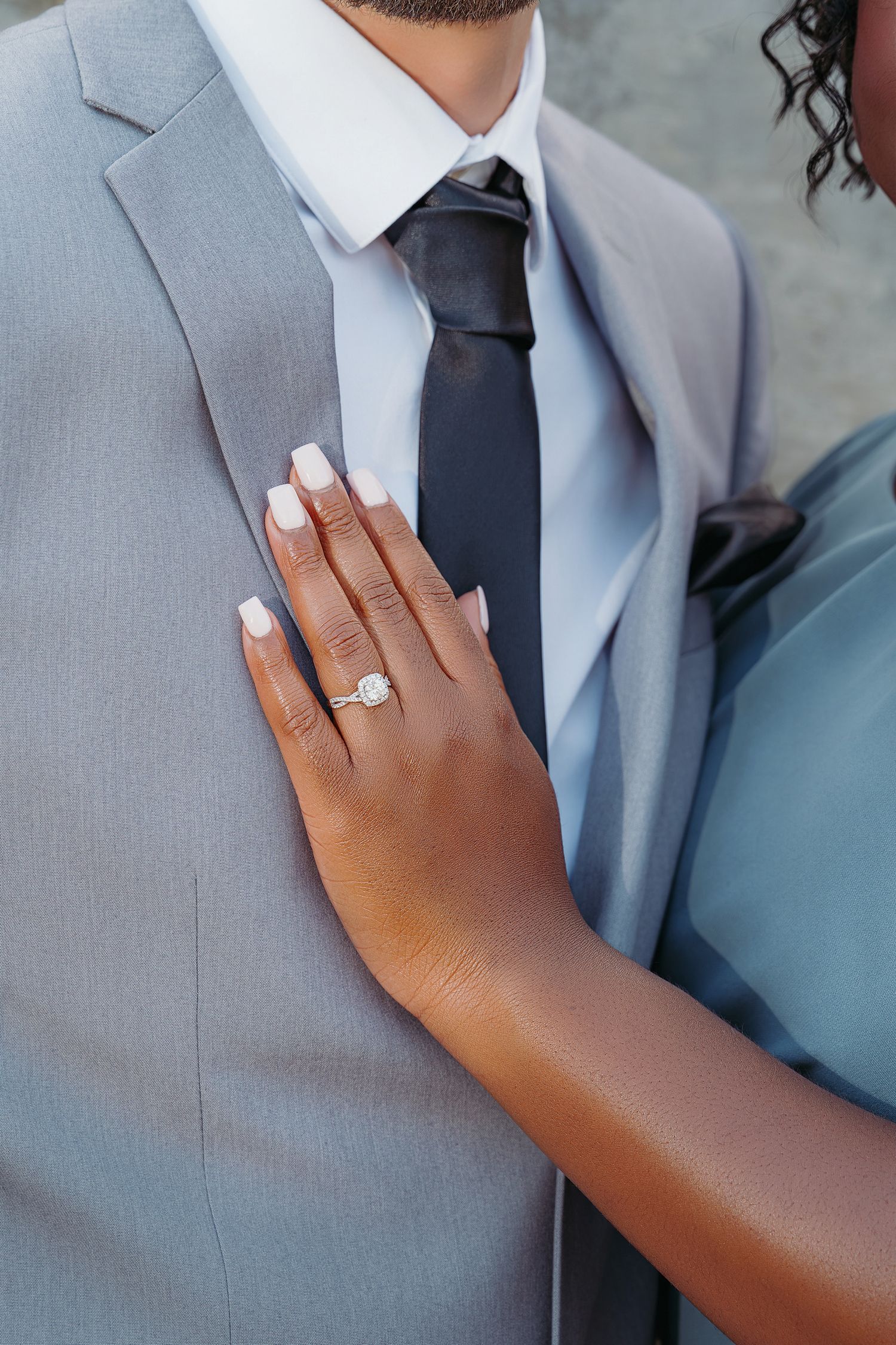 A hand with white manicured nails and a diamond engagement ring rests on a grey suit with black tie.