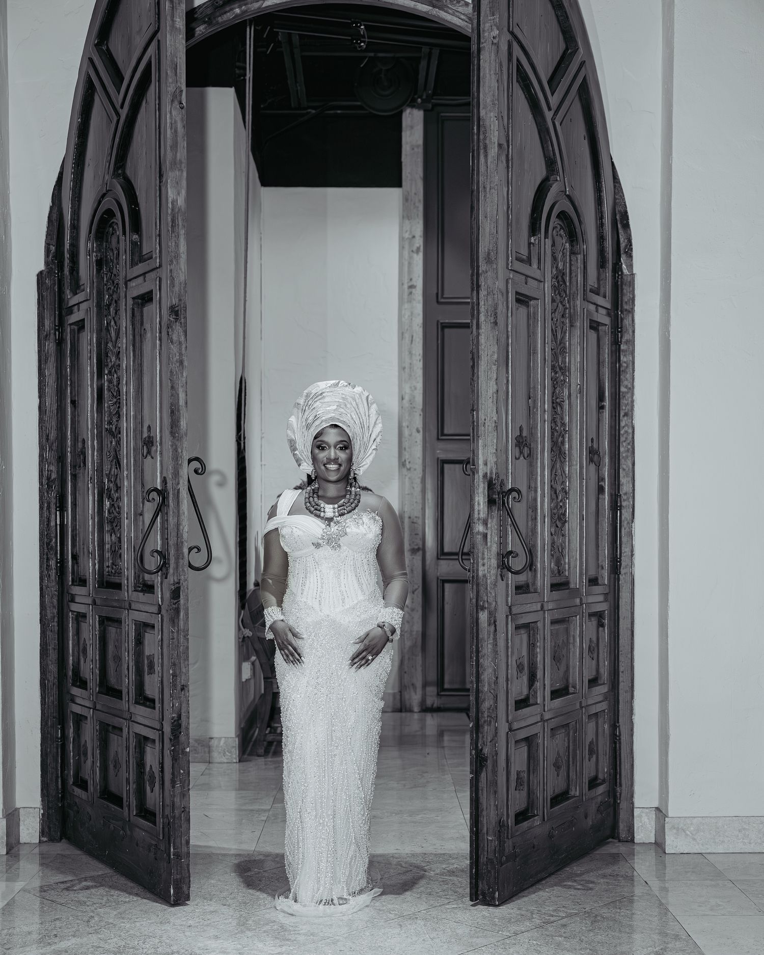 A figure in an elegant white dress and headpiece stands between ornate arched wooden doors in a black and white photograph.