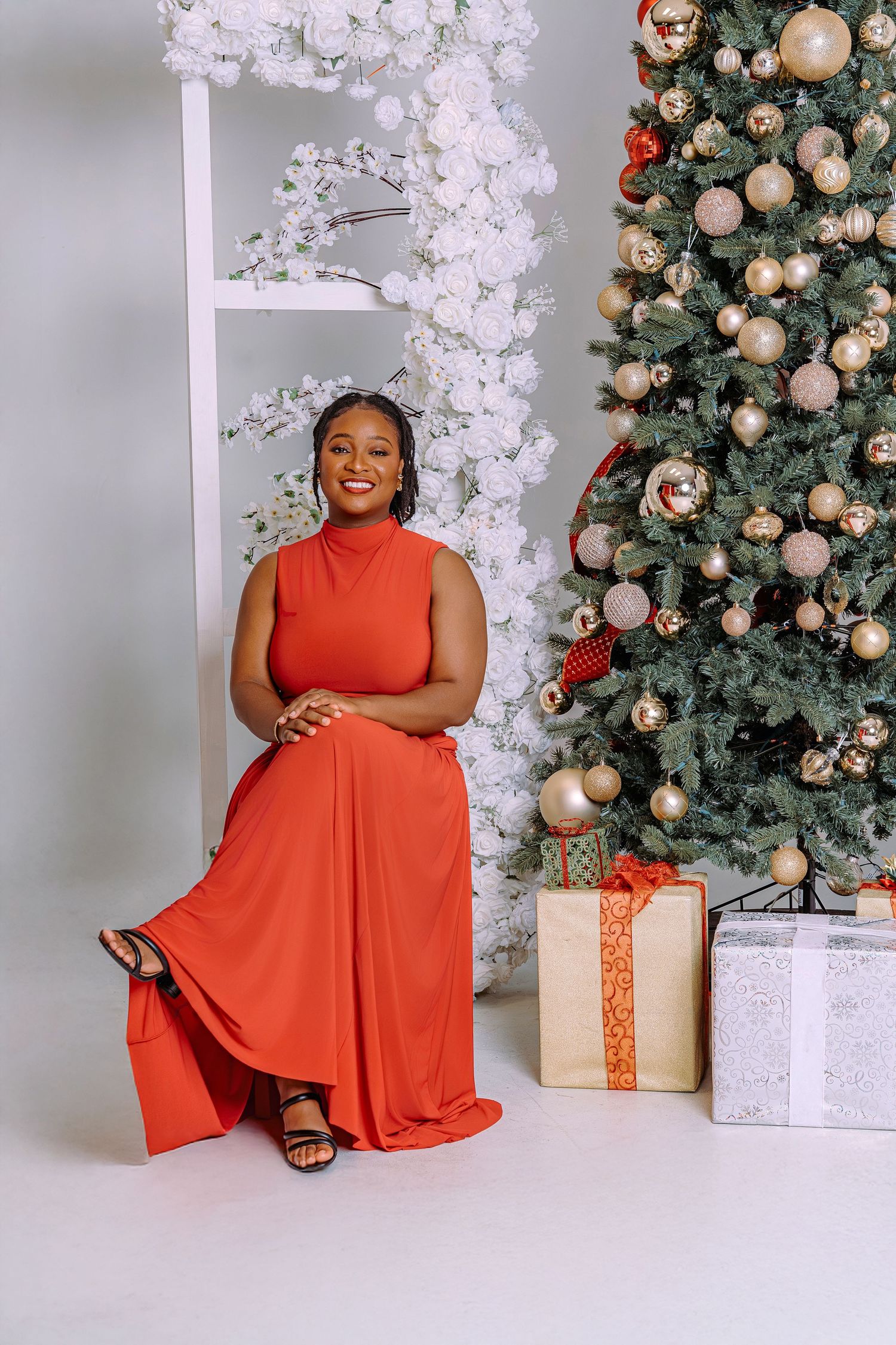 Woman in red dress smiling beside decorated Christmas tree with gold ornaments and wrapped gifts in white studio.