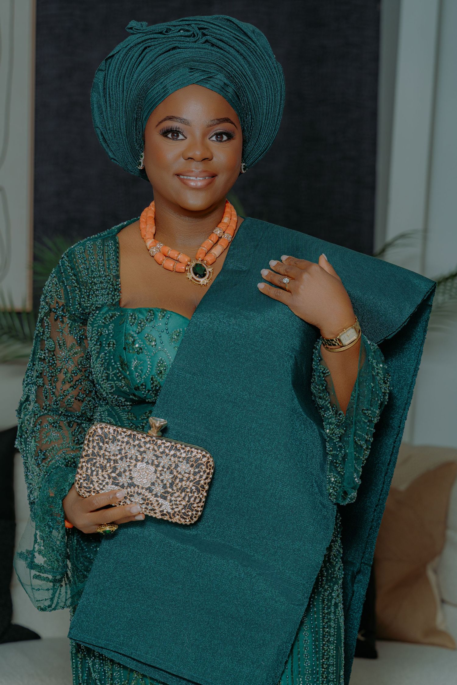 Woman in teal Nigerian traditional attire with gele headwrap, coral beaded necklace, and embellished clutch purse.
