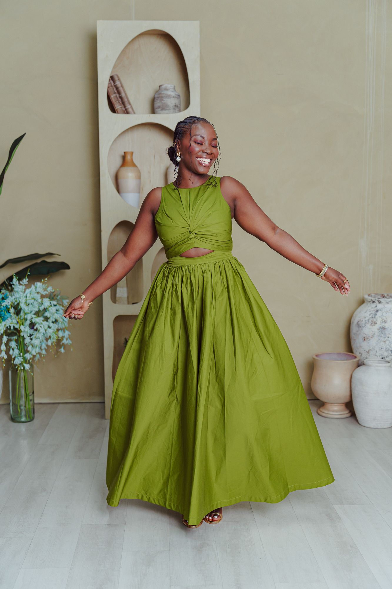 A green cotton dress with a full skirt is shown in multiple poses against a neutral backdrop with vases and flowers.
