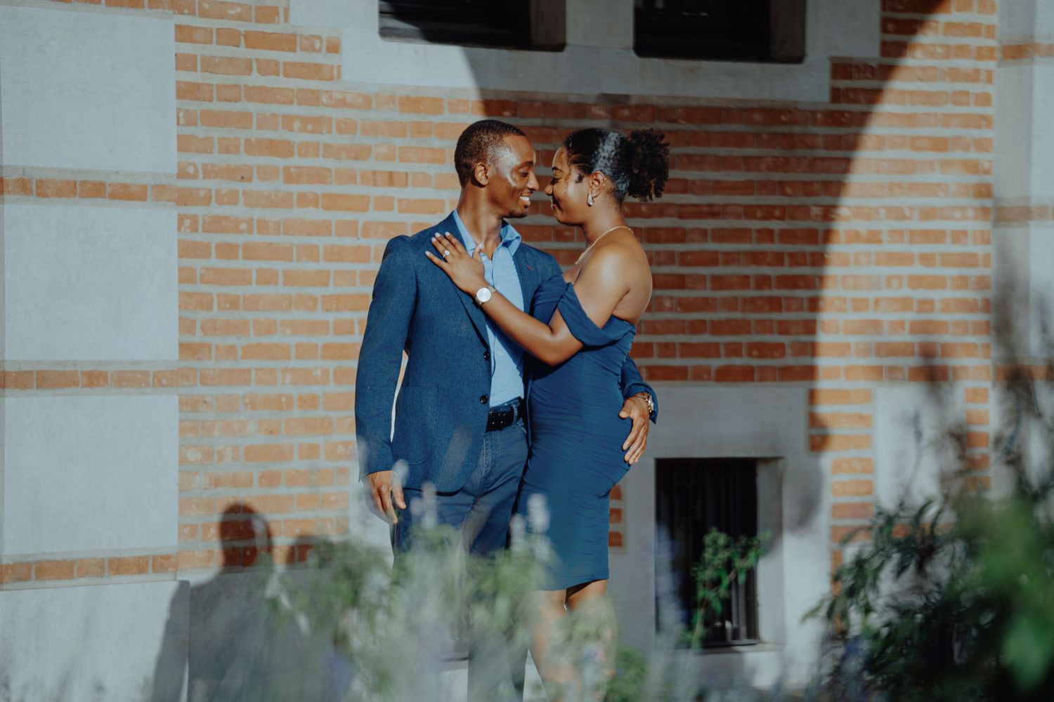A couple in formal attire shares a romantic moment outdoors near a brick building surrounded by greenery.