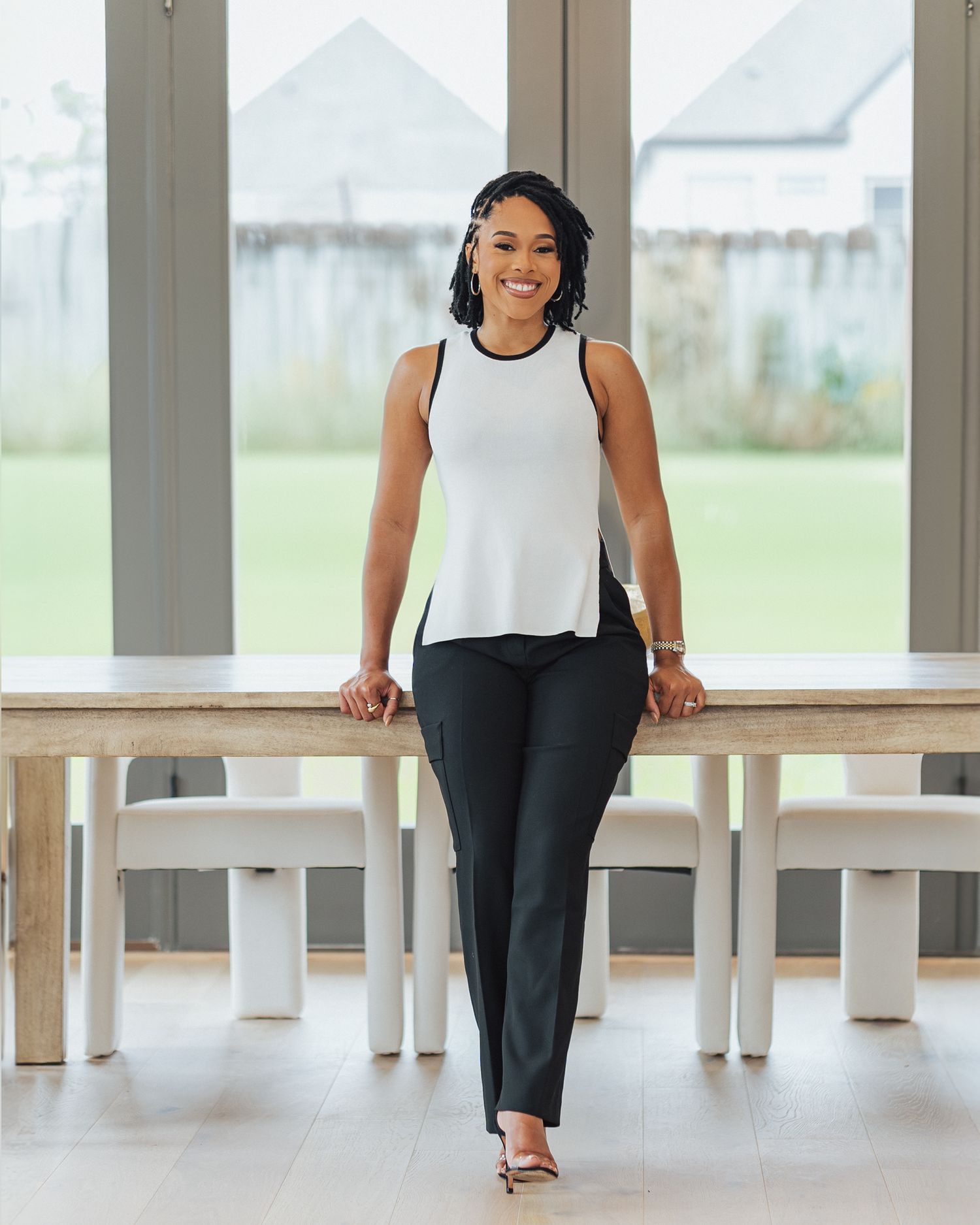 Smiling woman in white sleeveless top and black pants leaning against a wooden table in a bright modern room.