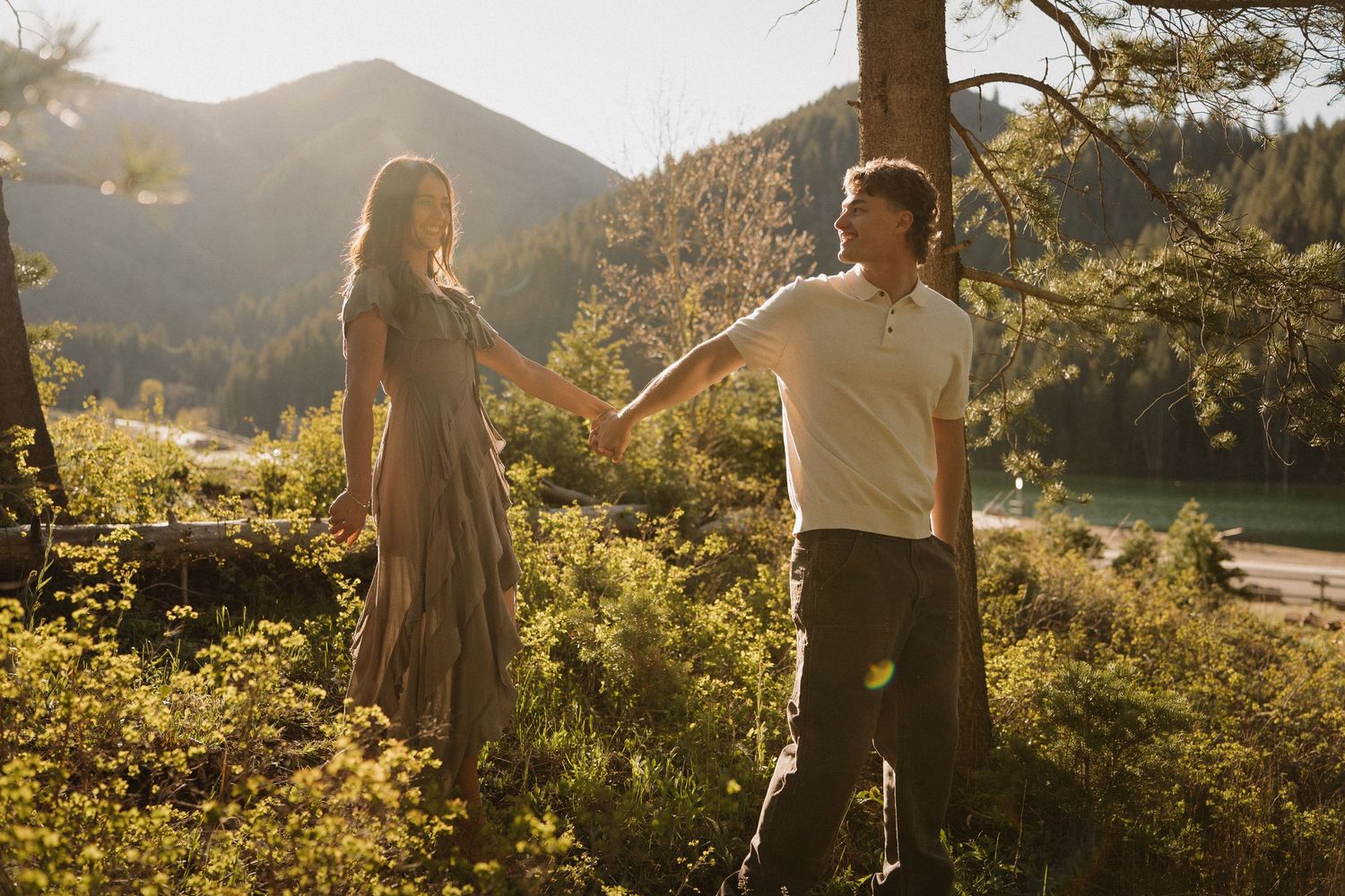 Couple holding hands in golden sunlight among lush greenery with mountain and lake backdrop.