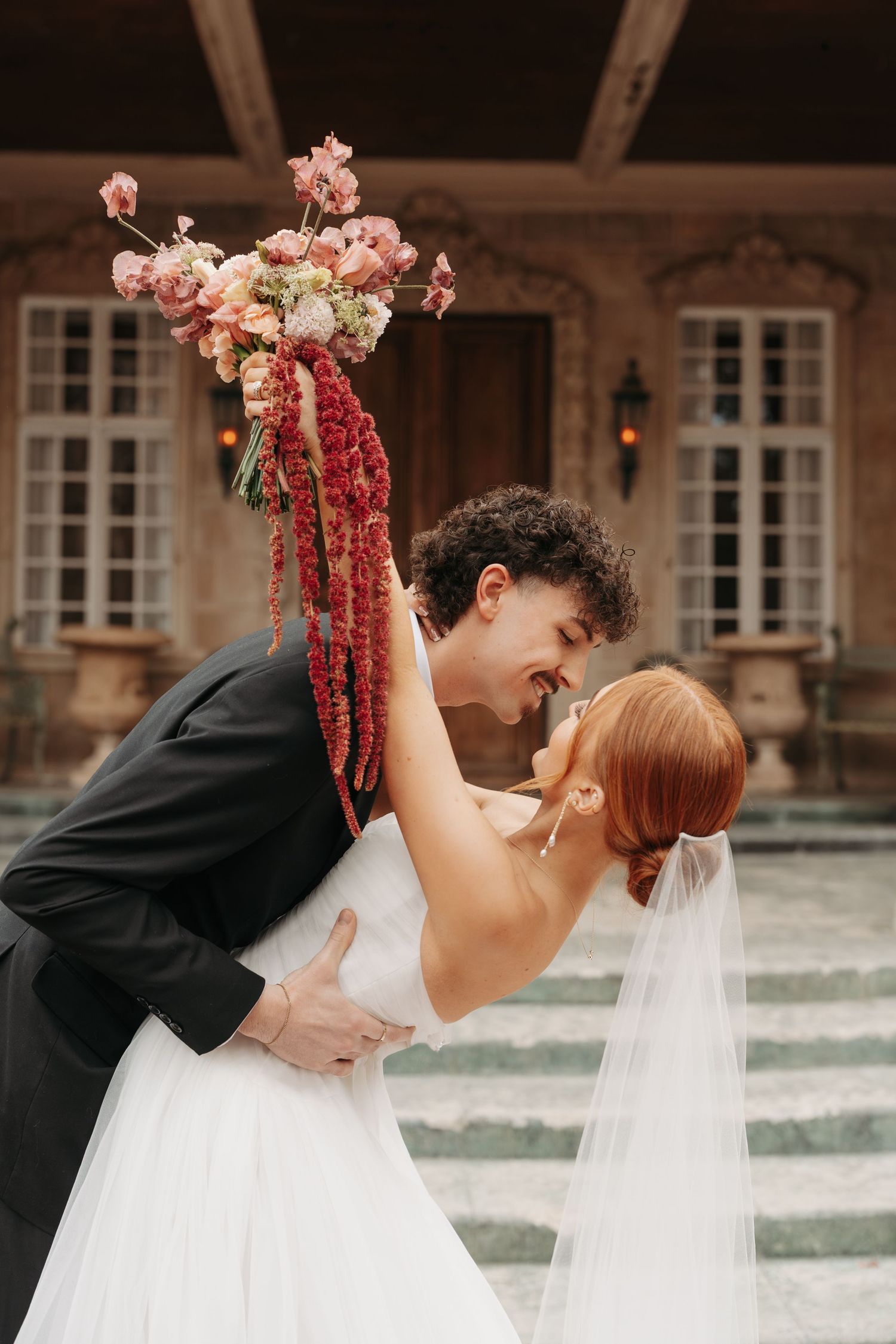 Romantic wedding couple dips and embraces while holding a pink floral bouquet in front of an elegant mansion.