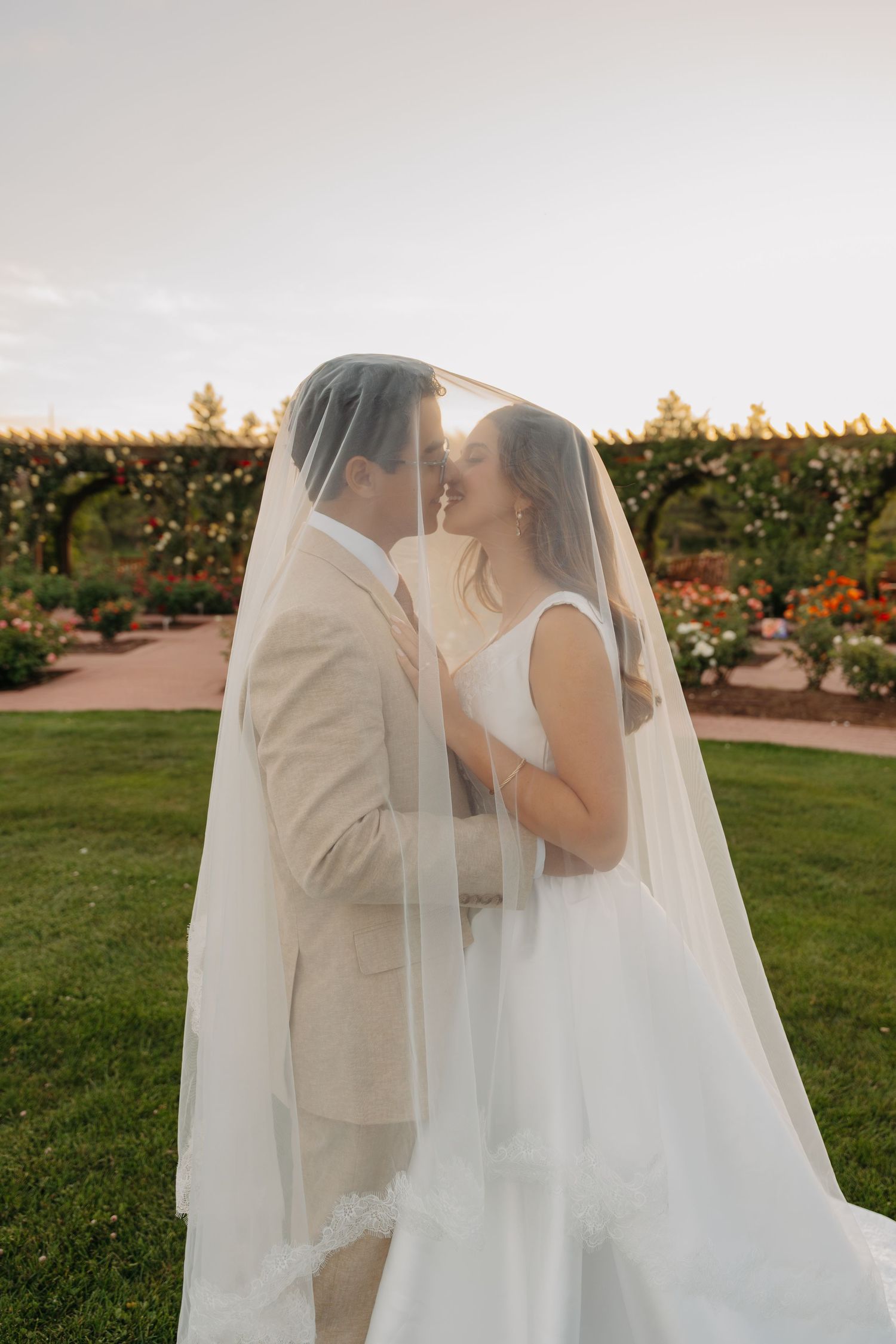 Bride and groom share a romantic moment under a flowing veil in a lush garden at sunset.