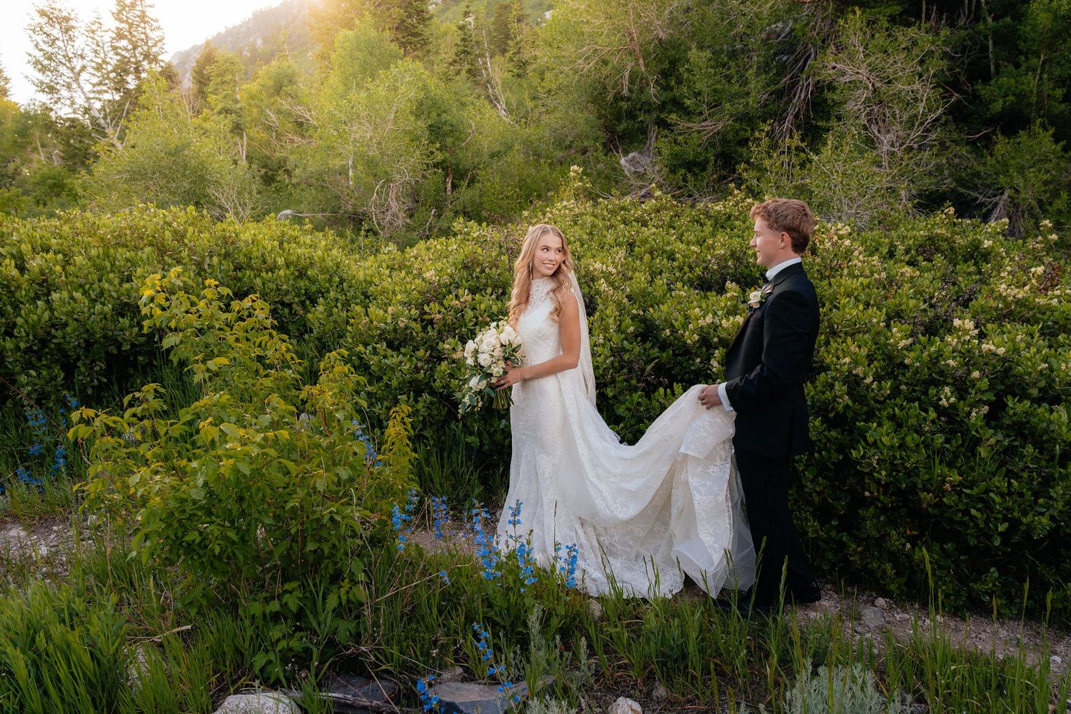 Bride and groom walk through lush green mountain landscape at golden hour, bride holding white bouquet in flowing gown.