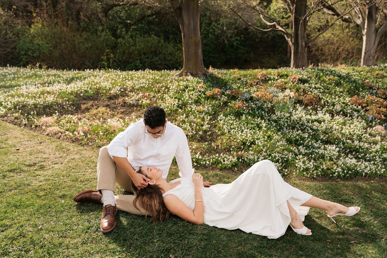 Couple in white attire relaxing on green grass near blooming spring flowers in a romantic outdoor setting.