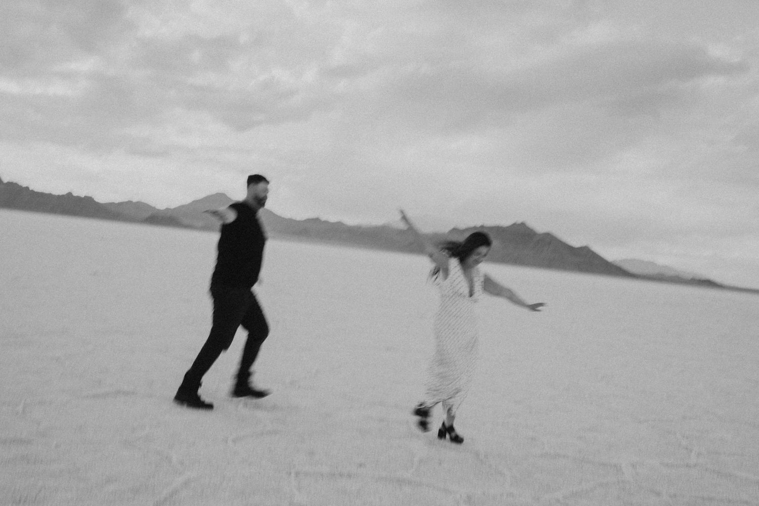 Black and white photo of couple dancing playfully on vast salt flats with dramatic cloudy sky.