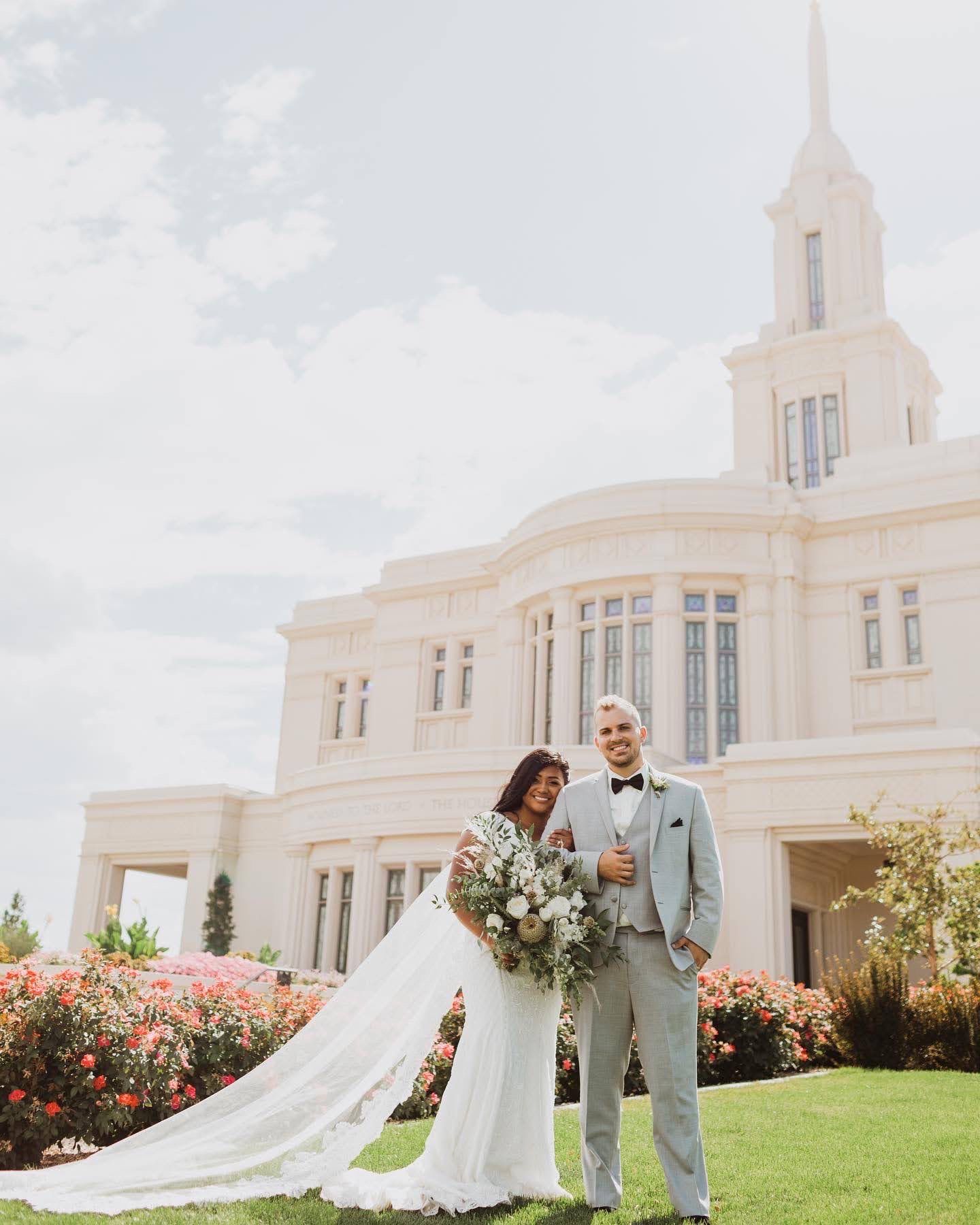 Wedding couple poses on lawn in front of majestic white temple building with floral bouquet.