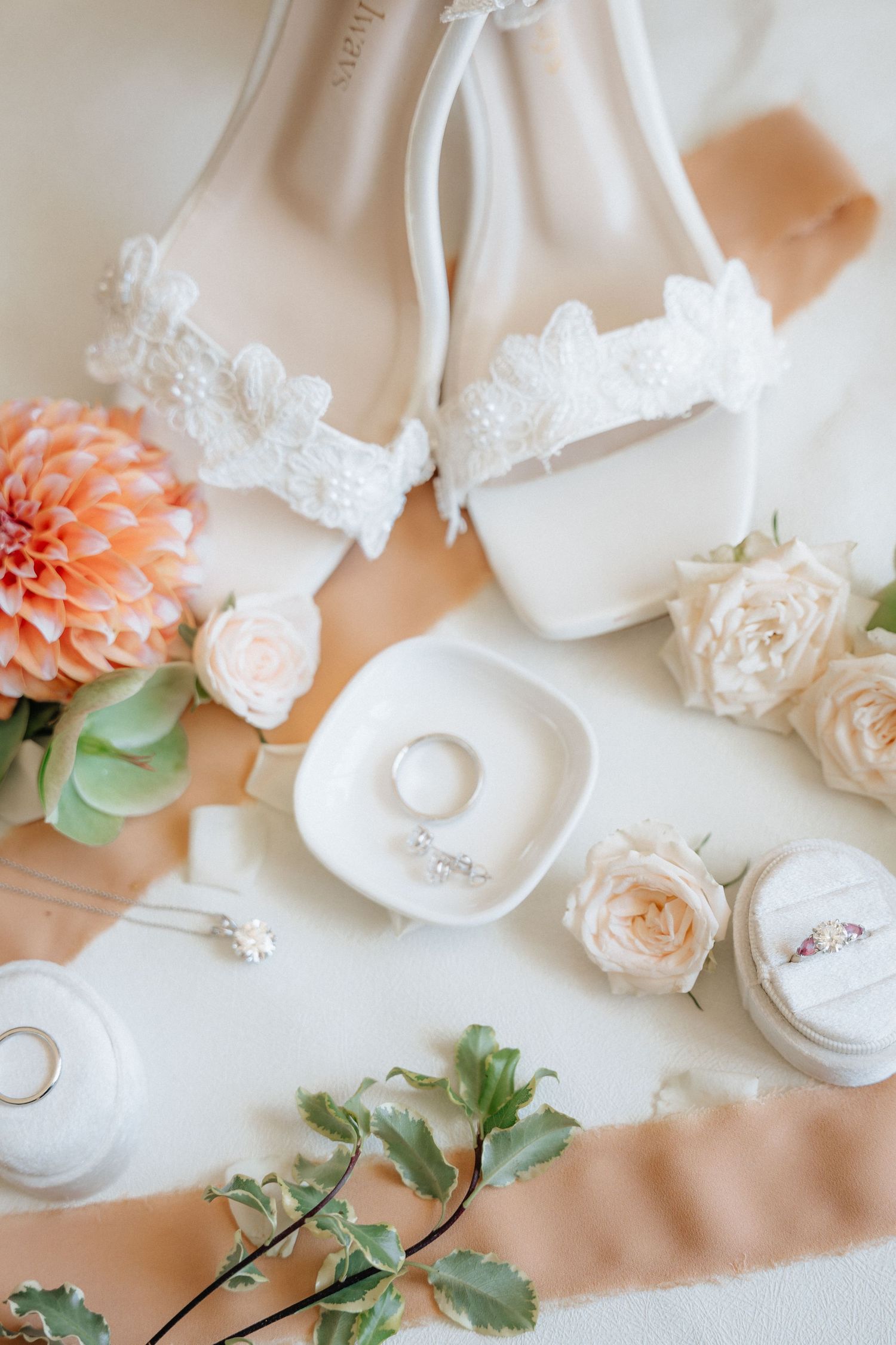 Elegant bridal flat lay with white lace heels, diamond rings, blush roses, peach dahlia, and satin ribbon on white background.