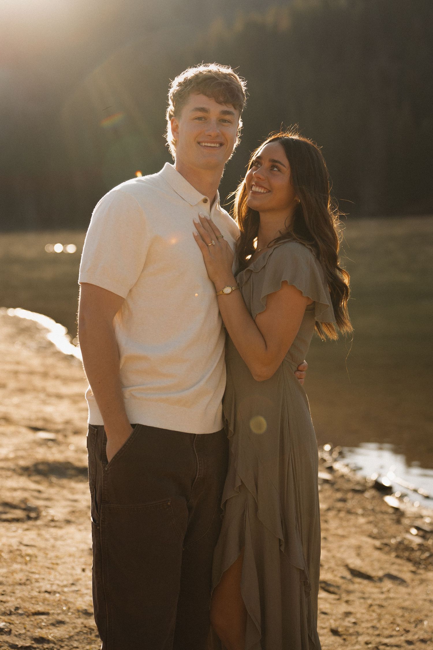 Couple posing at golden hour by a lake, woman in olive dress smiling up at man in white shirt.