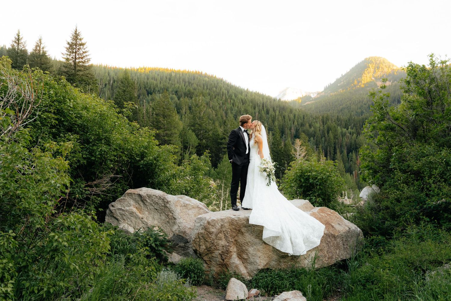 Bride and groom standing on a boulder surrounded by lush green forest and mountain scenery at sunset.