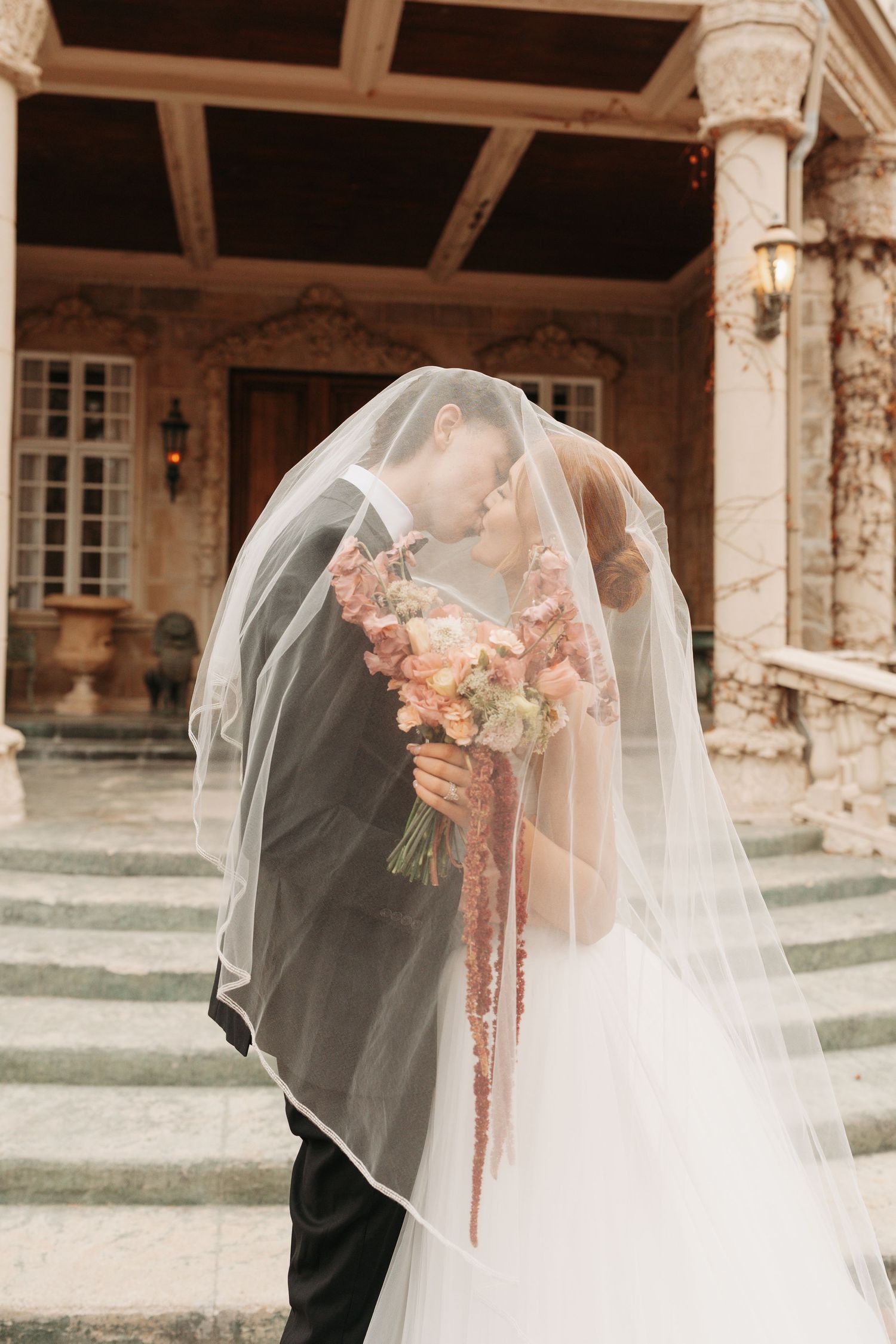 Elegant wedding couple shares an intimate moment under a stone archway with flowing veil and pink bouquet.