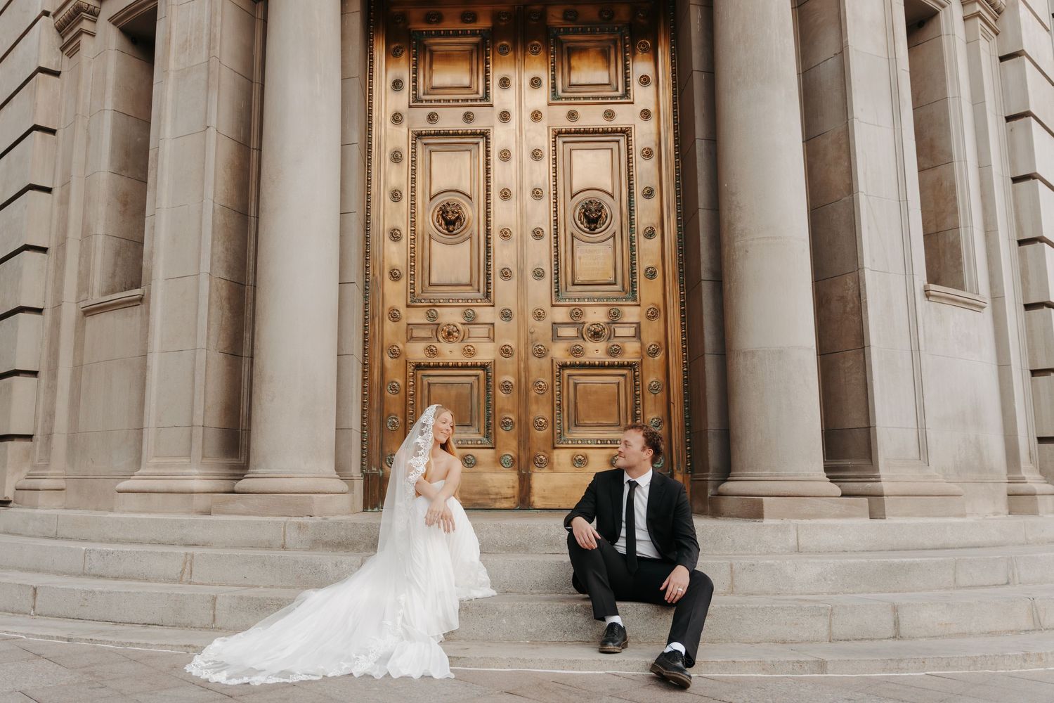 A wedding couple stands in front of ornate stone columns and wooden doors at a grand cathedral entrance.