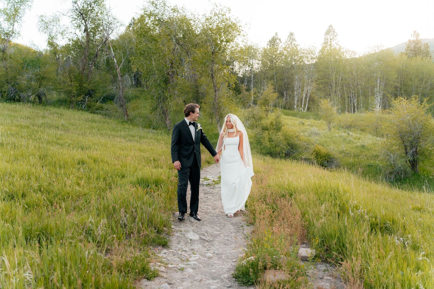 Bride and groom walk hand in hand along a scenic path through lush green meadow at golden hour.