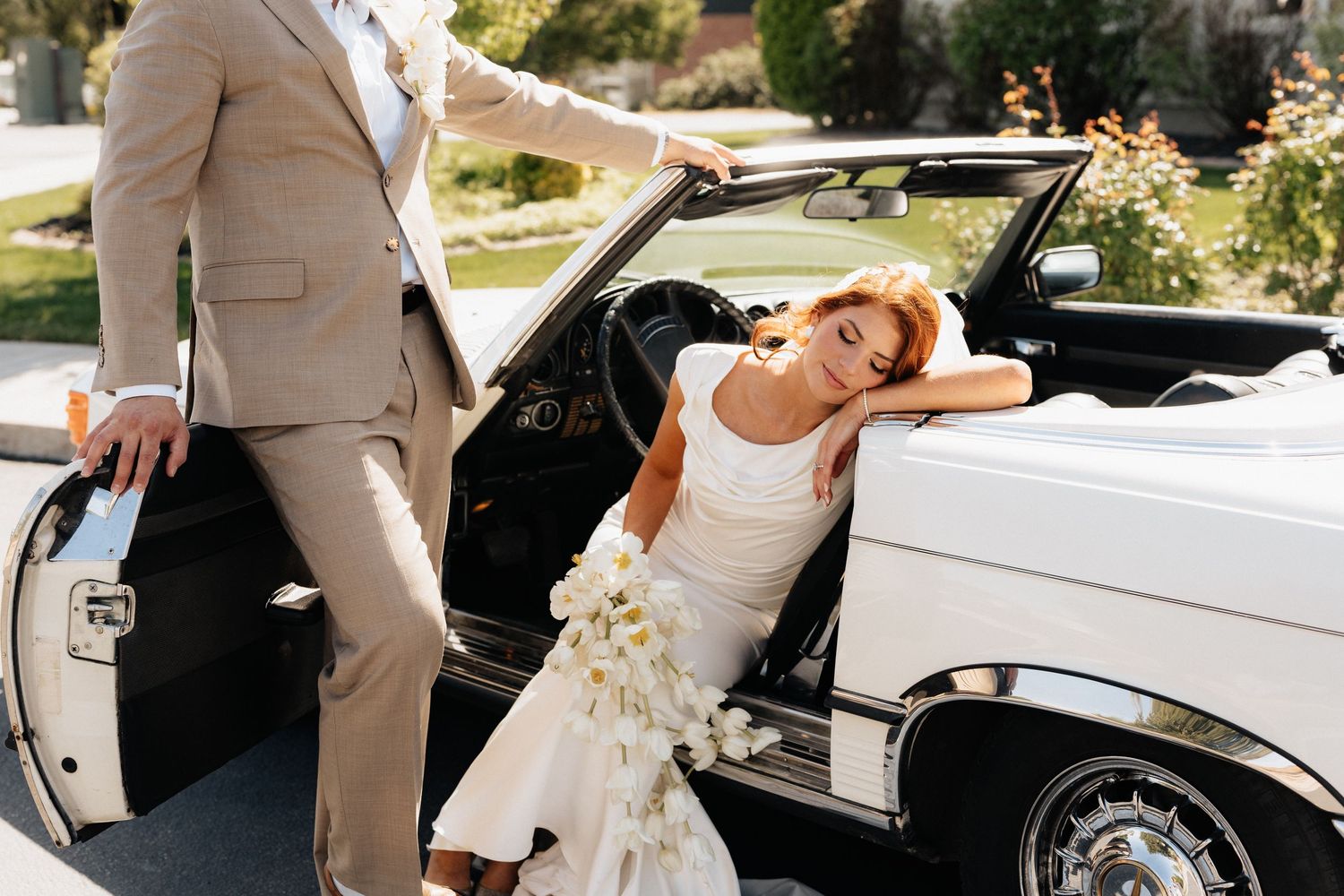 Bride in white gown exits vintage white convertible as groom in tan suit holds door open outdoors.