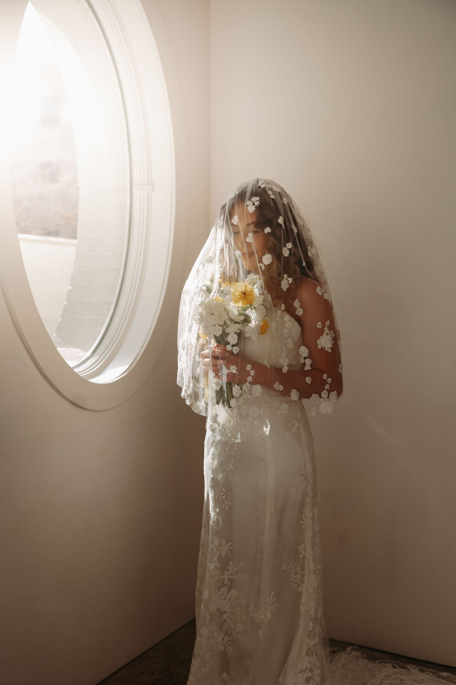 A wedding dress with floral details is displayed in front of a circular window in a minimalist white room.