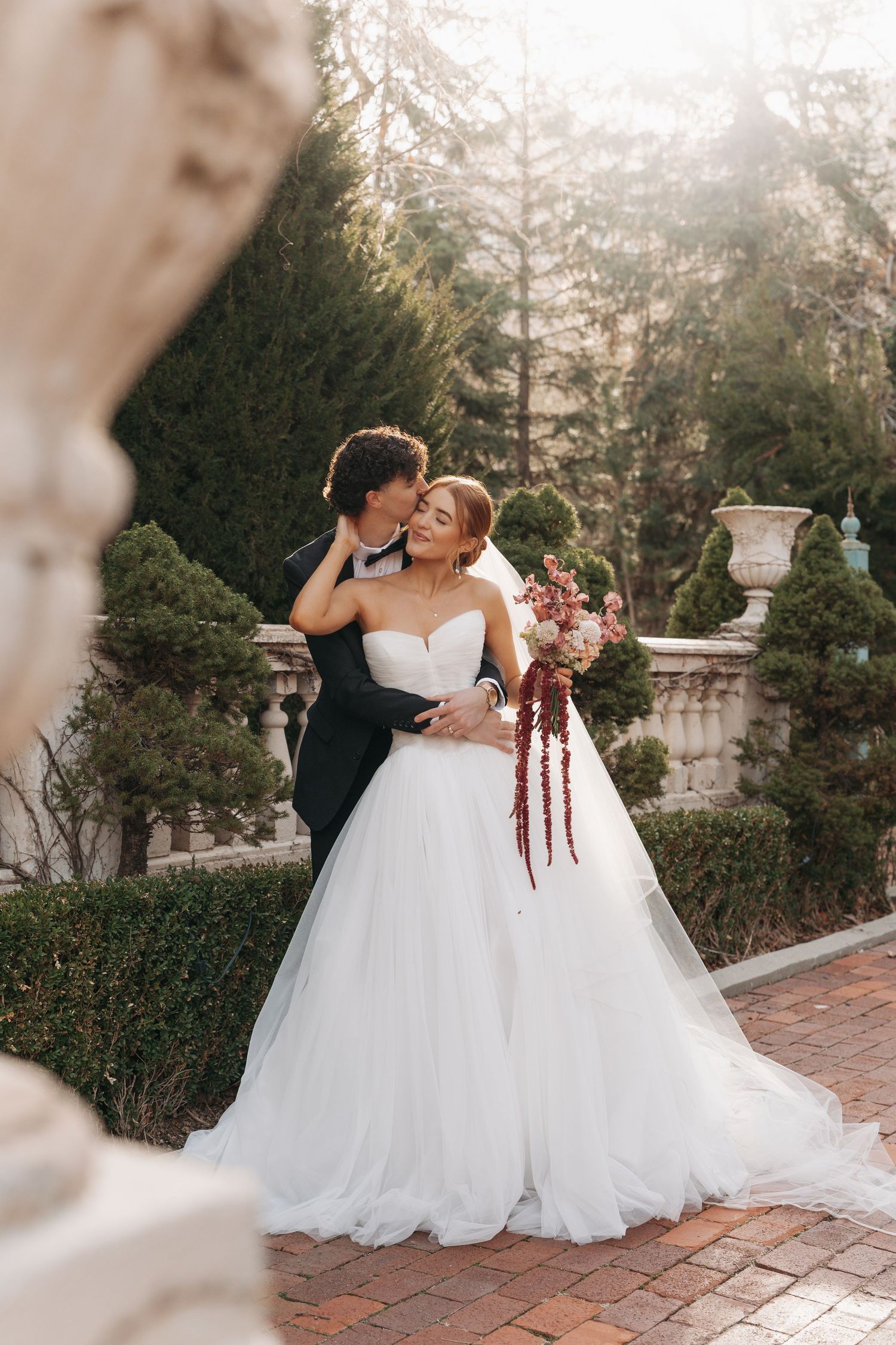 Bride in flowing white gown and groom embrace on brick pathway surrounded by manicured garden scenery.
