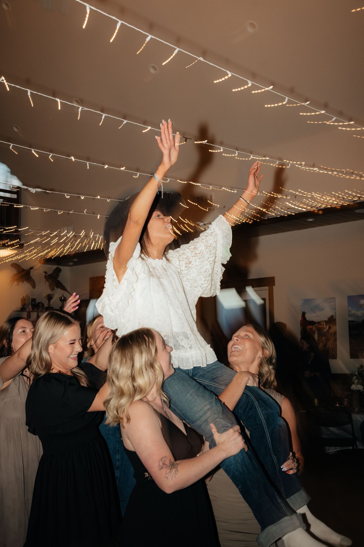 A bride in white is lifted by friends on a dance floor adorned with twinkling fairy lights at a wedding reception.